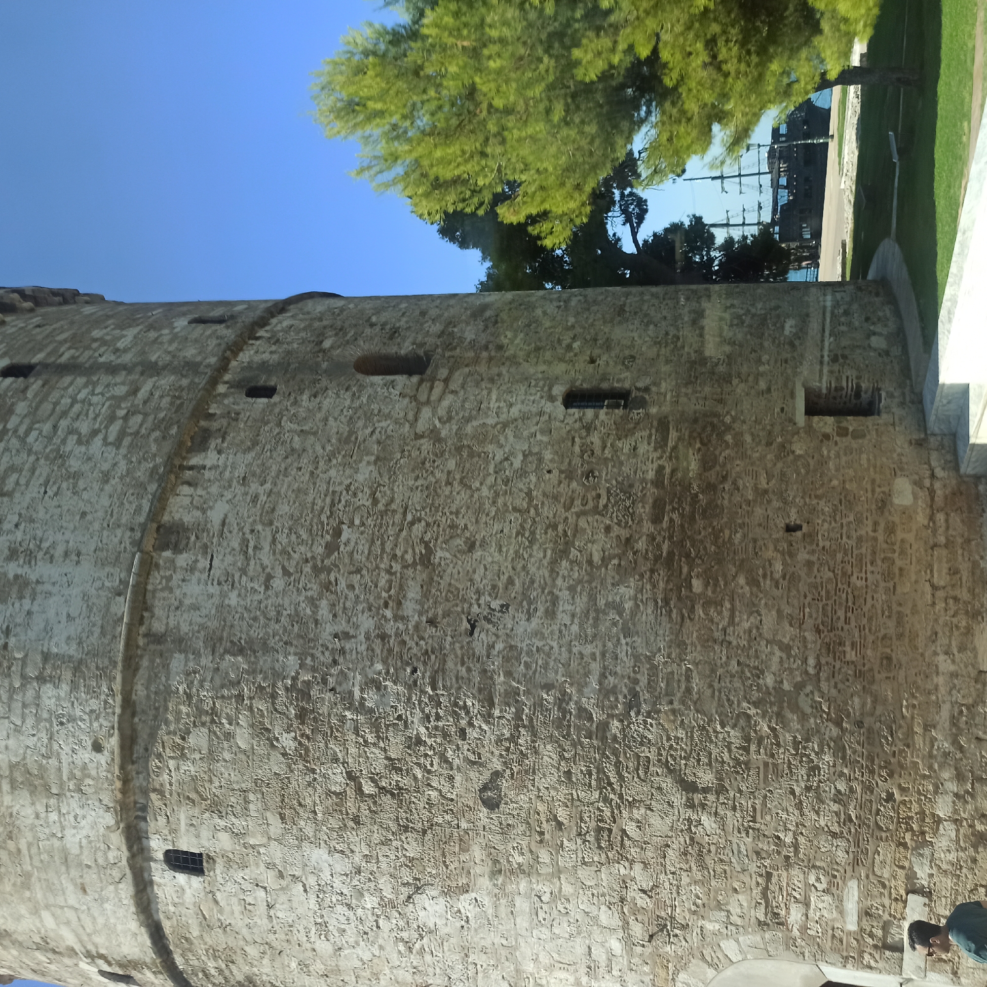The image displays the White Tower, a prominent landmark in Thessaloniki, Greece. The tower is a cylindrical stone structure with visible brickwork and small, dark openings that serve as windows. A man wearing sunglasses and a dark shirt is partially visible in the lower left corner of the frame. In the background, a lush green tree occupies the right side of the image, and behind it, a tall-masted ship is docked in the blue water, with a clear blue sky overhead. The foreground features a paved path and a patch of green grass.