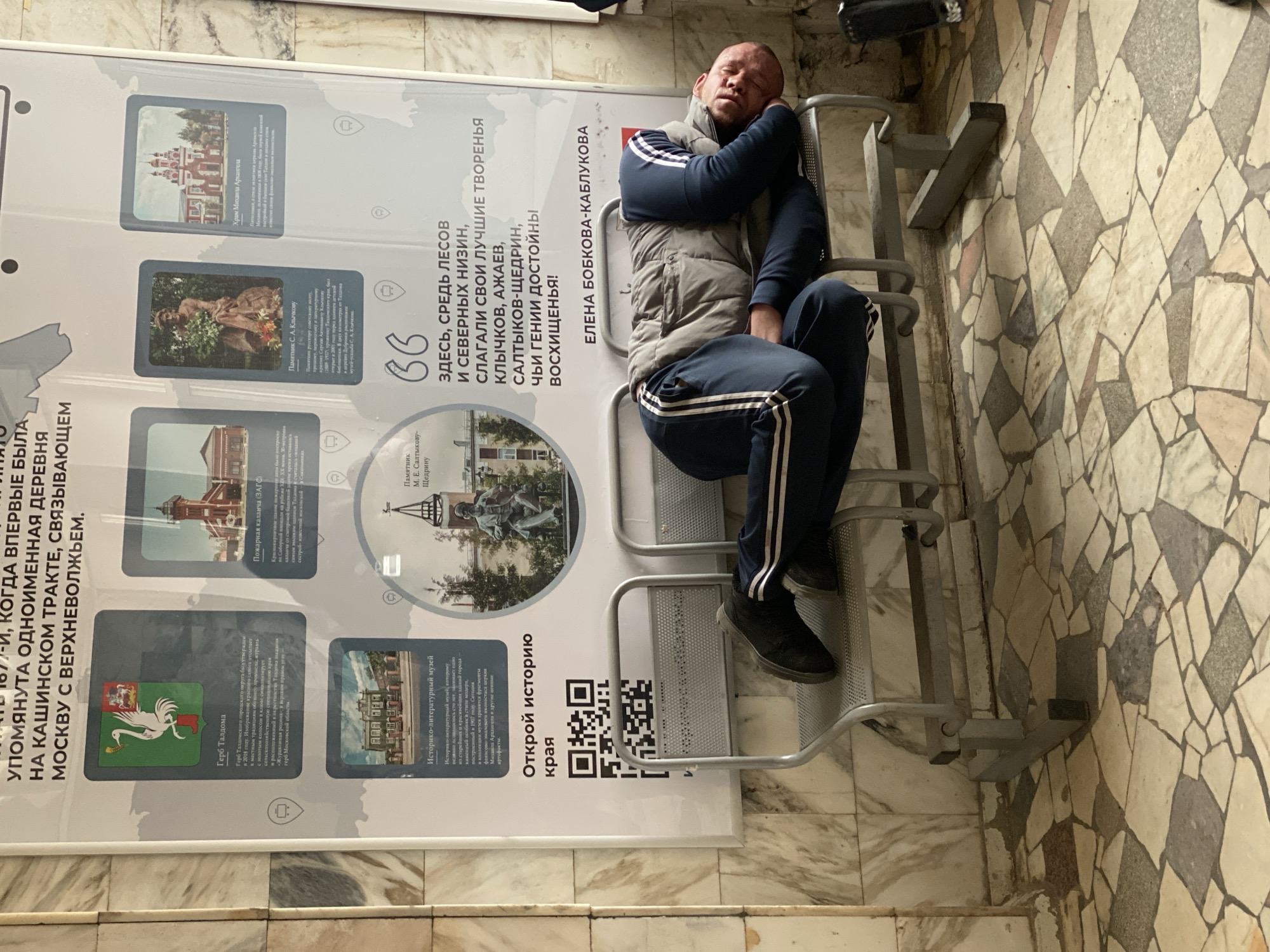 An indoor, eye-level shot captures a man resting on a metal waiting bench next to a large informational display. The man, with short hair and stubble, wears a grey puffer vest over a blue long-sleeved shirt with white stripes on the sleeves, and matching blue sweatpants with white stripes. He is lying on his side on the bench, with his head resting on his left arm, appearing to be asleep or deeply relaxed.
The floor is covered in a mosaic pattern of light-colored, irregular tiles, predominantly off-white, grey, and light brown. Behind the man is a tall, white informational stand or banner, displaying various historical and cultural points of interest, consistent with the provided location context of Taldom, Russia.
Visible text on the banner includes:
- A large quote: "ЗДЕСЬ, СРЕДЬ ЛЕСОВ И СЕВЕРНЫХ НИЗИН, СЛАГАЛИ СВОИ ЛУЧШИЕ ТВОРЕНЬЯ КЛЫЧКОВ, АЖАЕВ, САЛТЫКОВ-ЩЕДРИН, ЧЬИ ГЕНИИ ДОСТОЙНЫ ВОСХИЩЕНЬЯ!" (HERE, AMONG FORESTS AND NORTHERN LOWLANDS, KLYCHKOV, AZHAEV, SALTYKOV-SHCHEDRIN CREATED THEIR BEST WORKS, WHOSE GENIUSES ARE WORTHY OF ADMIRATION!).
- Another name: "ЕЛЕНА БОБКОВА-КАБЛУКОВА".
- Titles for featured images: "Памятник С. А. Клычкову" (Monument to S. A. Klychkov), "Храм Михаила Архангела" (Michael the Archangel Church), "Герб Талдома" (Coat of Arms of Taldom), "Пожарная каланча (ЗАГС)" (Fire tower (ZAGS)), "Историко-литературный музей" (Historical and Literary Museum), and "Памятник М. Е. Салтыкову-Щедрину" (Monument to M. E. Saltykov-Shchedrin).
- A QR code with the text "Открой историю края" (Discover the history of the region).
- Text detailing the history of the Taldom coat of arms and other local facts.
The artificial lighting suggests it could be day or night. The overall atmosphere is quiet, possibly a waiting area where one might rest during travel.