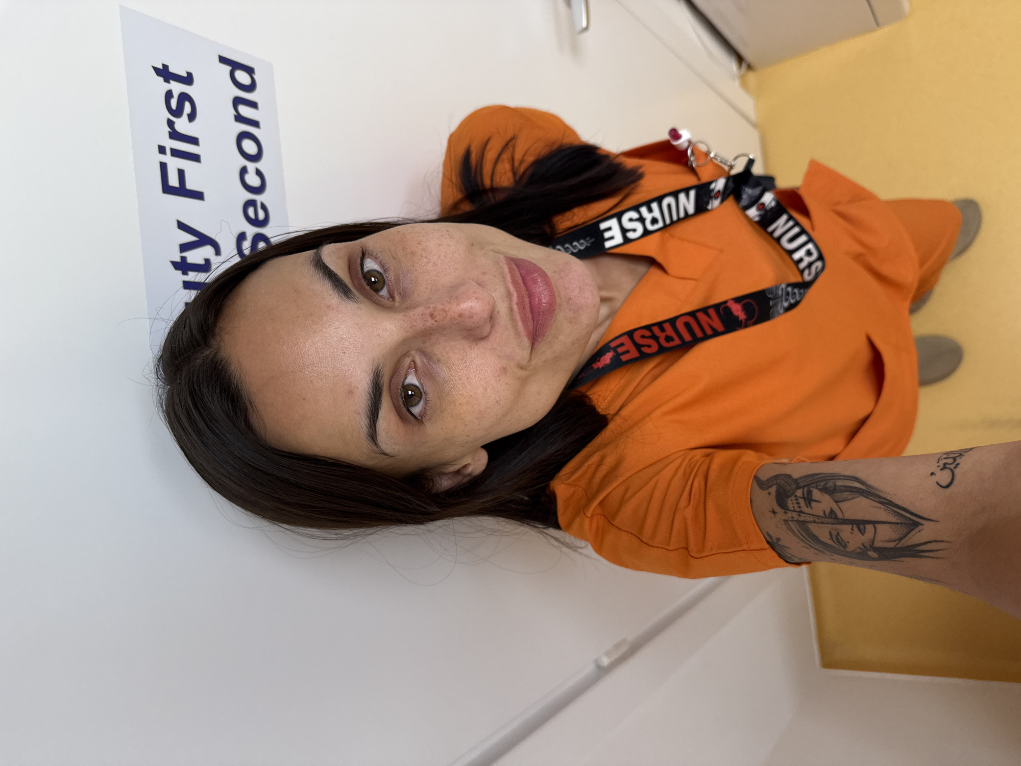 The image shows a close-up, selfie-style photograph of a woman wearing an orange scrub top and scrub pants. She has long dark hair and is looking at the camera with a slight smile. She has a tattoo on her left arm. A lanyard with the word "NURSE" printed on it is visible around her neck. In the background, a sign on the wall reads "Beauty First Second". The floor is yellow. The image is undated and shows no specific geographical indicators of the city of Pleven, Bulgaria.