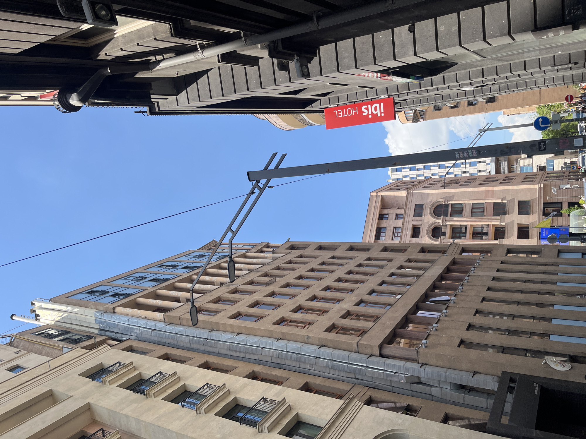 This is an upward-angled view of a bustling urban street in Yerevan, Armenia, captured on a bright, sunny day. The scene is dominated by various architectural styles of tall buildings and overhead structures.
In the upper left, the dark, ribbed underside of an elevated structure or building overhang is prominent, featuring visible pipes and lighting fixtures. A bright red sign with white text, clearly reading "ibis HOTEL", hangs prominently below this overhang.
Several multi-story buildings rise into the clear blue sky, which has scattered white clouds. A large, light brown building with numerous square windows and some small balconies stands prominently on the left. Next to it, a taller building features modern, reflective glass windows. Further in the background, another building with distinct arched windows can be seen.
A street lamp arm extends from a building facade on the left. In the distance, a pole with a circular blue traffic sign indicates a left-turn arrow. Faint glimpses of street-level activity, including vehicles and people, are visible further down the street on the right. The strong sunlight and clear sky suggest it is likely midday or early afternoon.