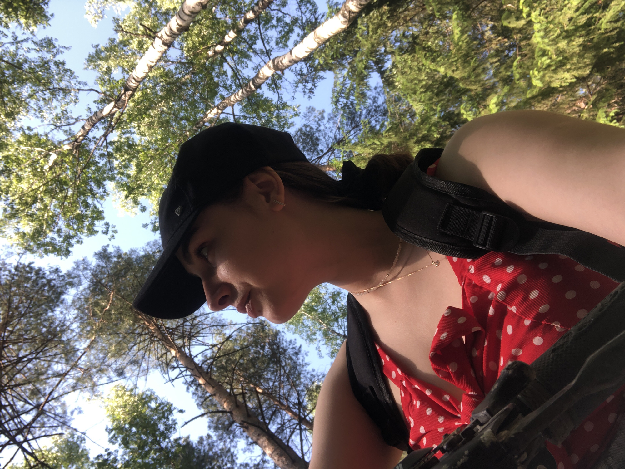 A woman wearing a black baseball cap and a red and white polka dot dress stands in a forest. She is looking down and to the left. She is wearing a black shoulder strap. The trees are green and the sky is blue.