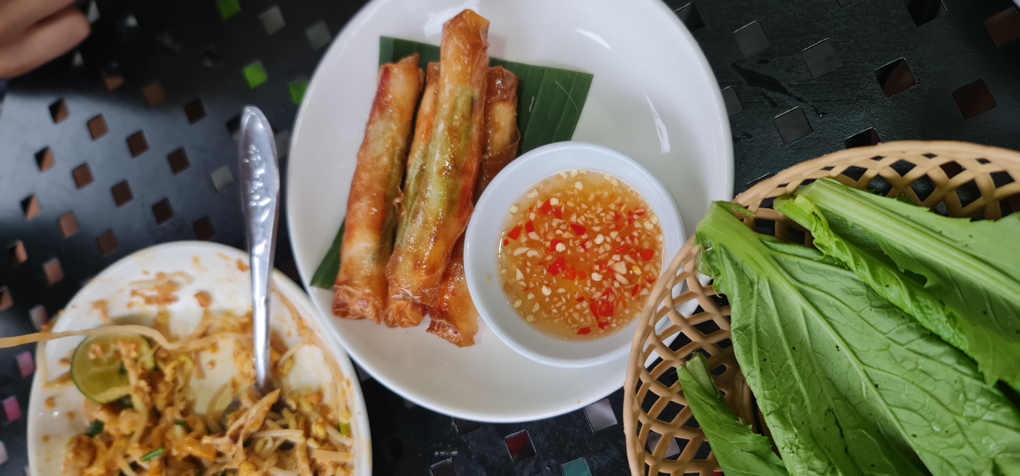 The image shows a top-down view of a table setting with dishes, potentially in Hội An Ward, Vietnam. The table has a dark surface with a geometric square pattern. On the table, there is a white plate containing several spring rolls arranged on a green leaf, next to a small bowl filled with a dipping sauce that contains visible red chili pieces. Adjacent to the plate is another white dish with leftover noodles, possibly Pad Thai, and a utensil resting on the side of the dish. A woven basket filled with green leafy vegetables is located to the right of the plate with the spring rolls. The composition suggests a casual dining setting with a mix of appetizers, a main course, and fresh greens.