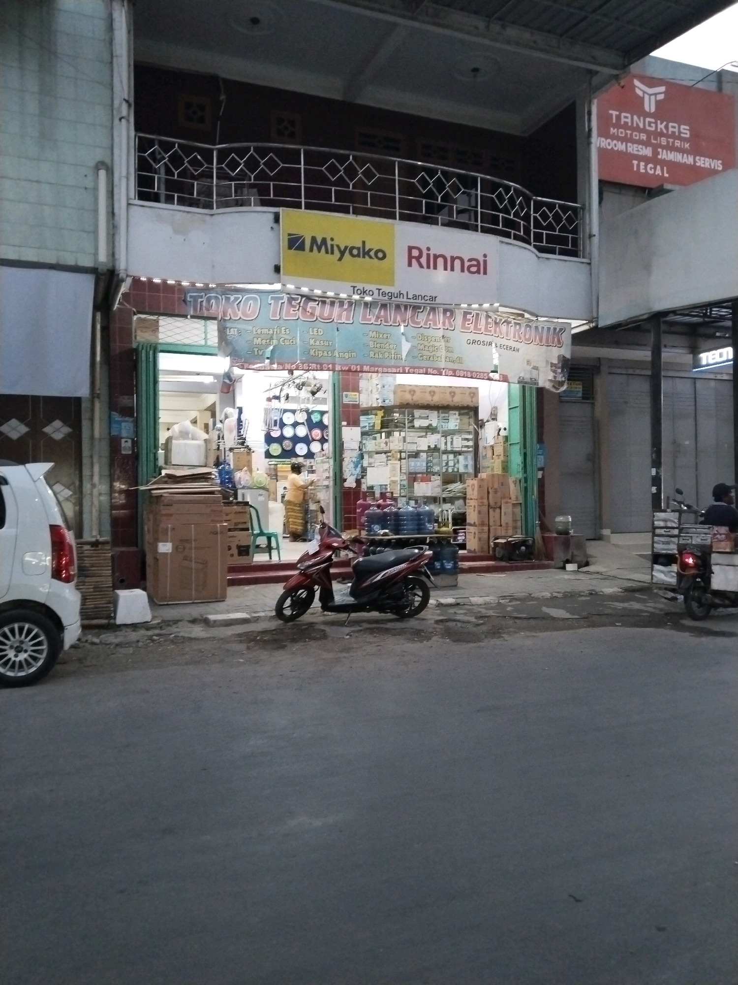 The image captures a street view in front of an electronics store in Margasari, Indonesia. The store, named "TOKO TEGUH LANCAR ELEKTRONIK," has a prominent sign displaying the brands "Miyako" and "Rinnai." The storefront is open, revealing shelves stocked with various electronic appliances and goods. A woman in traditional Indonesian attire is visible inside the store. In front of the store, several boxes and a green chair are placed on the sidewalk. A red scooter and a white car are parked on the street, with another scooter with a rider in the background. Adjacent to the electronics store, a sign for "TANGKAS MOTOR LISTRIK" is visible, indicating a motorcycle repair shop. The street is paved, and the overall scene suggests a typical commercial area in an Indonesian city.