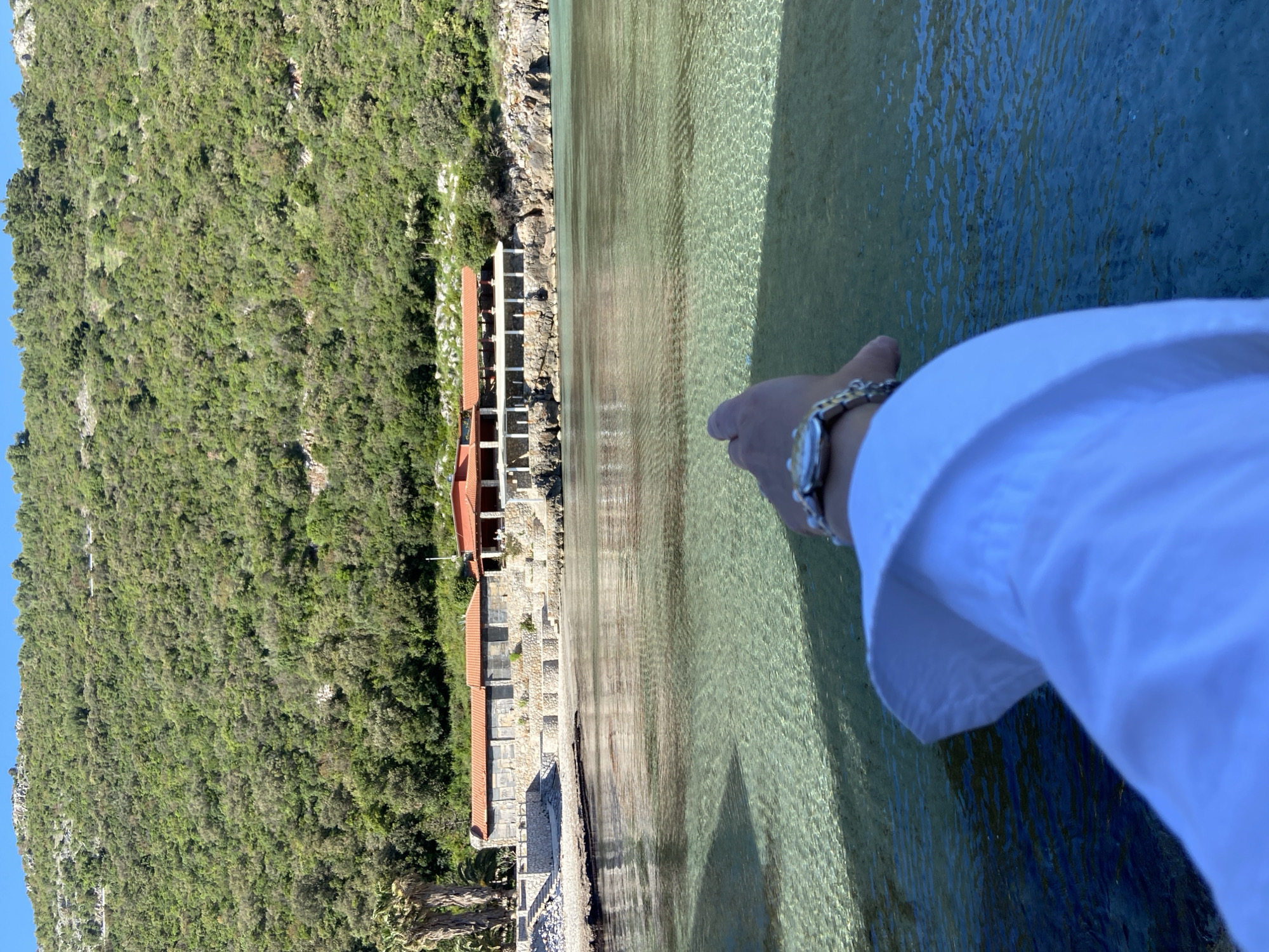 The image shows a view of a coastline with buildings and lush greenery, likely in Visnjeva, Montenegro, based on the prompt. The landscape features a steep, green hill on the left side, contrasted against a series of buildings with red-tiled roofs along the shoreline. The water in the foreground appears clear with visible ripples. In the right foreground, a person’s arm wearing a silver watch and a white shirt sleeve is visible. The focus of the image seems to be the buildings and the natural surroundings, suggesting a scenic shot of the coastal area.