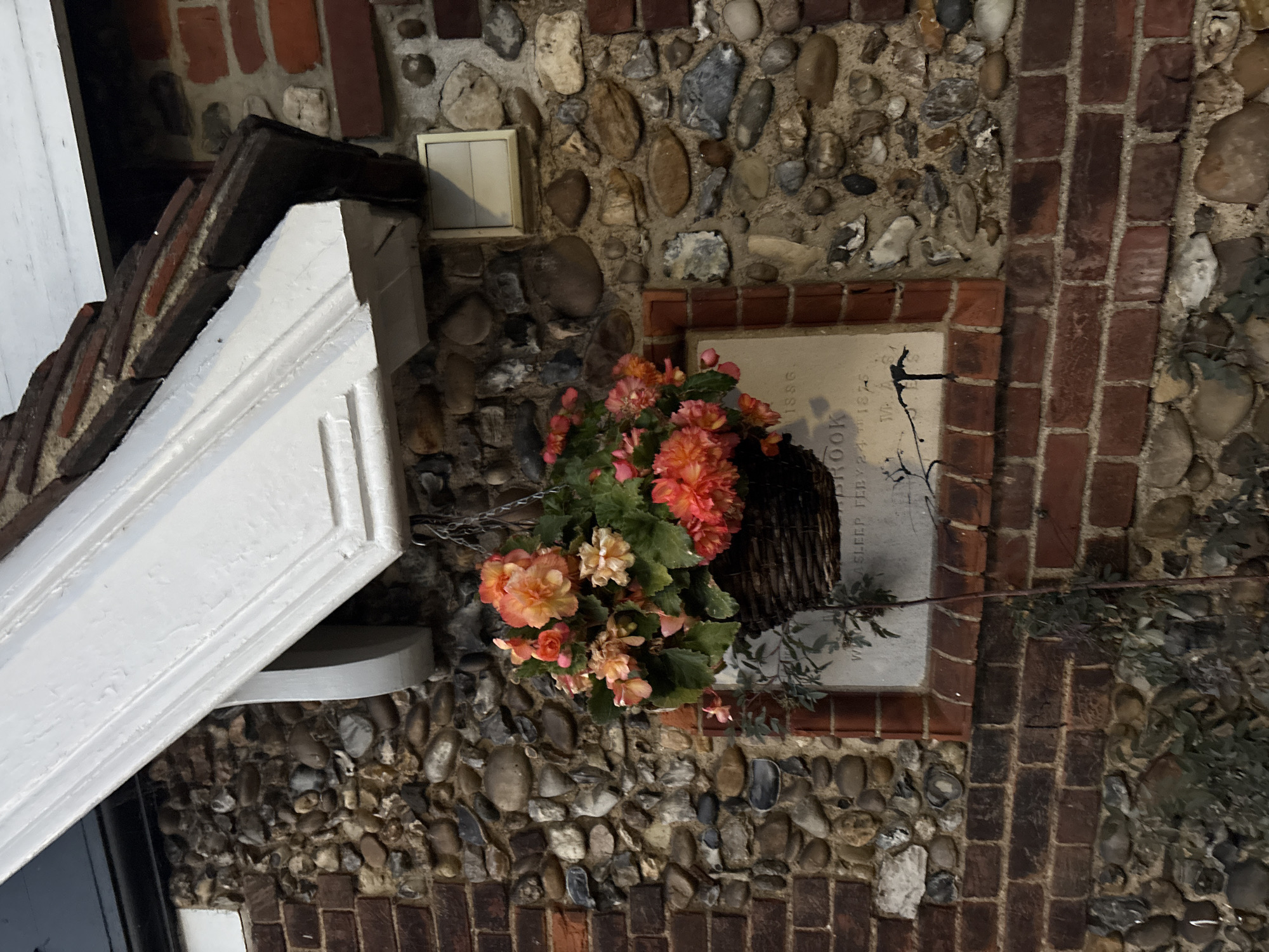 The image shows a hanging basket filled with orange and pink begonia flowers, suspended from a chain against a rustic stone and brick wall. A white painted canopy extends from the roofline above, casting a shadow. Below the hanging basket, a plaque is visible, partially obscured by the foliage. The plaque appears to have an inscription that includes the year "1886" and other text. The wall is constructed of various sized stones and red bricks. The overall scene suggests an outdoor environment, possibly in a historical setting within the city of Chelmsford, United Kingdom.