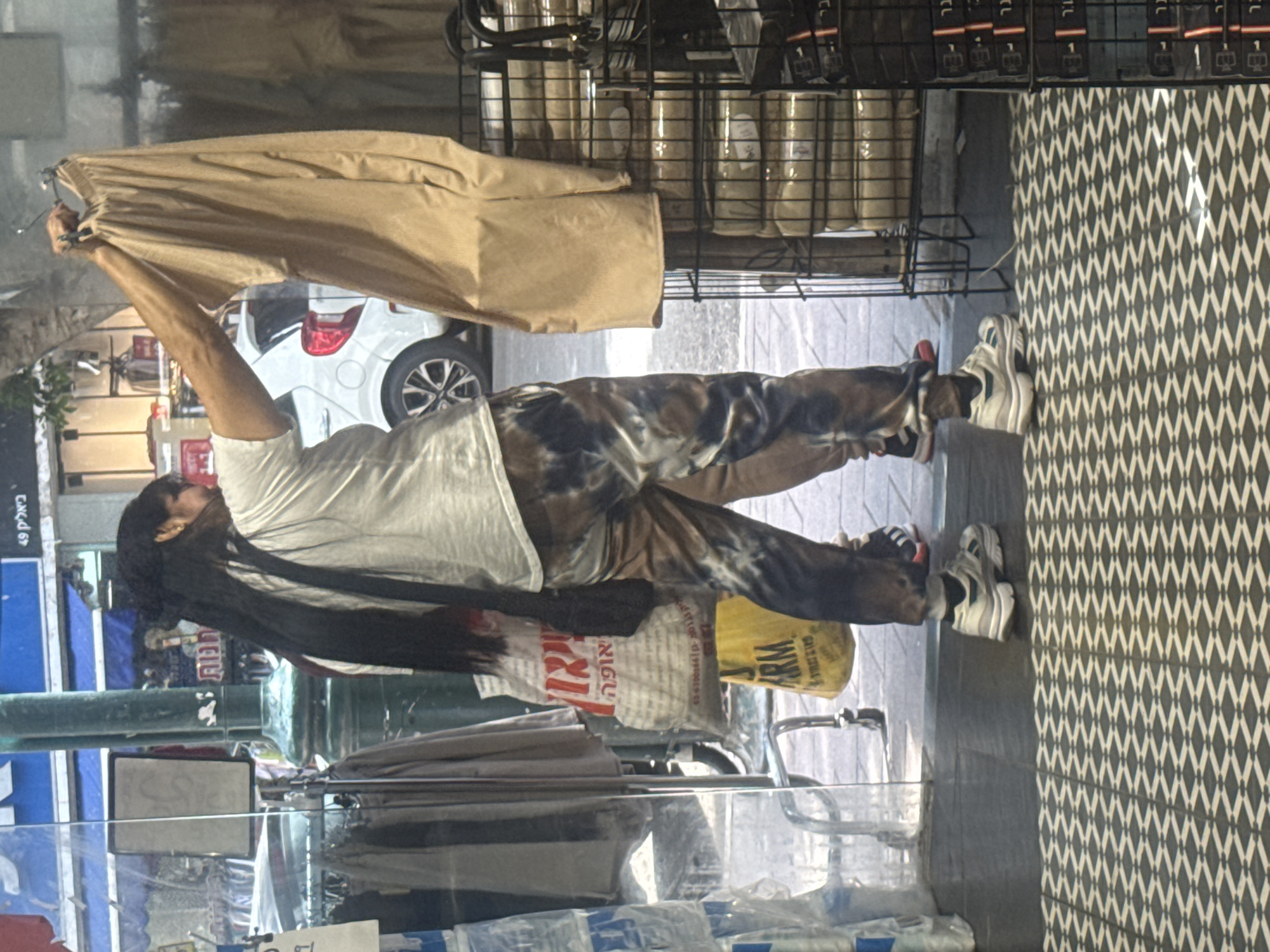 A woman is standing inside a store in Ramat Gan, Israel. She is holding a pair of khaki pants above her head. She is wearing a white shirt and tie-dye pants, with white sneakers. She has long dark hair. There is a car parked outside the store. The store has a display case and metal shelving. The floor is tiled with a geometric pattern. There are shopping bags at her feet.