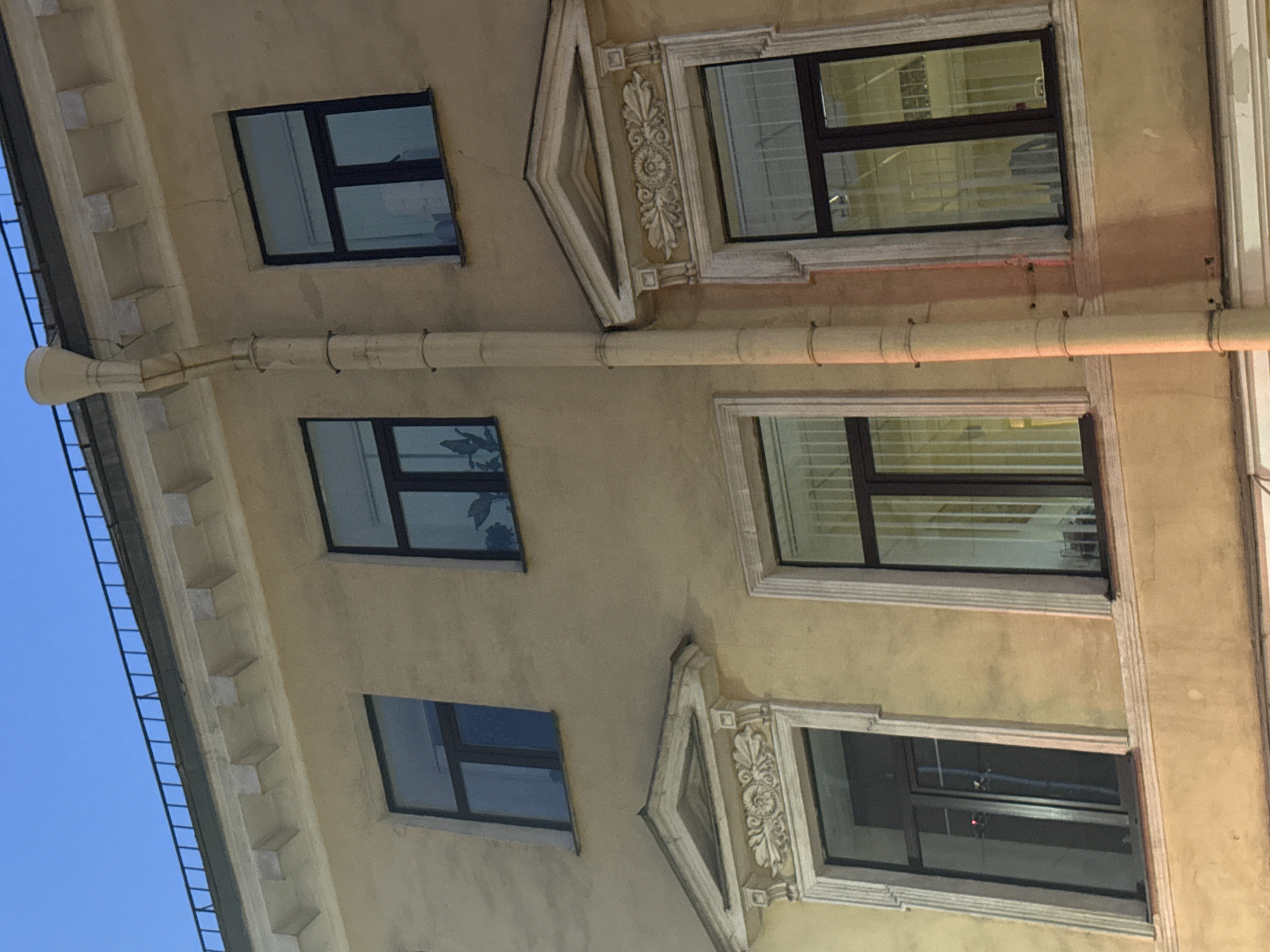 The image shows a facade of a building in Saint Petersburg, Russia. The building has a yellow stucco exterior and features several windows with dark frames. Some of the windows have decorative pediments above them. A downspout runs vertically along the side of the building. The sky above is a clear blue. The image is taken from a low angle, looking up at the building. There are no people visible in the image.
