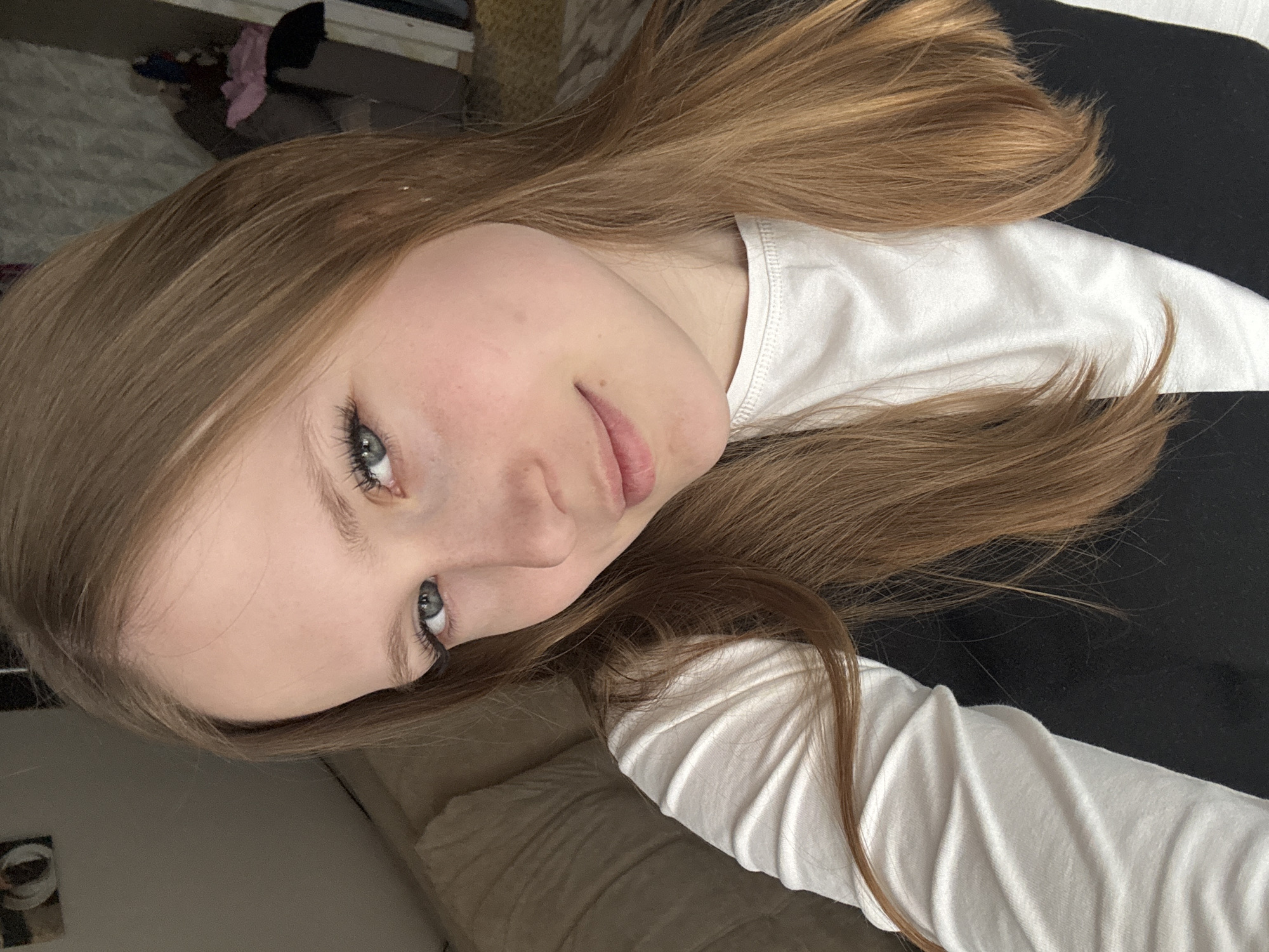 A close-up, tilted, eye-level shot shows a young woman with long, light brown hair and fair skin. She has light-colored eyes and a slight smile. She is wearing a white, long-sleeved shirt over a black garment. The background is indistinct, showing a textured beige surface and various objects in the far distance. The image was likely taken indoors. No specific location in Chaikovsky, Russia is identifiable.