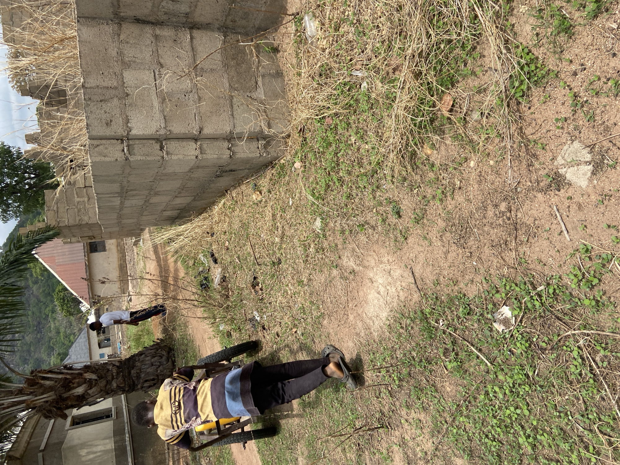 An outdoor, daytime scene depicts a semi-developed area, likely in Mararaba, Nigeria. The ground is a mix of bare dirt, dry, straw-like grass, and sparse patches of small green vegetation.
In the foreground, a person wearing a striped shirt and dark pants is seen from behind, seated or kneeling on a small, yellow-wheeled cart. A large palm tree trunk is prominent to their left. Further in the midground, another individual in a white t-shirt and dark pants stands on a dirt path, facing away from the camera.
A tall, partially constructed concrete block wall stretches across the left side of the scene. Beyond this wall, several modest buildings with tin roofs are visible. In the distant background, green, undulating hills rise under a bright sky, suggesting clear weather.
The dry state of the grass indicates a dry season or arid conditions. Some scattered litter, including a white plastic bottle, can be seen on the ground near the concrete wall. No specific activity beyond the presence of the individuals is clearly depicted, and there is no visible text.