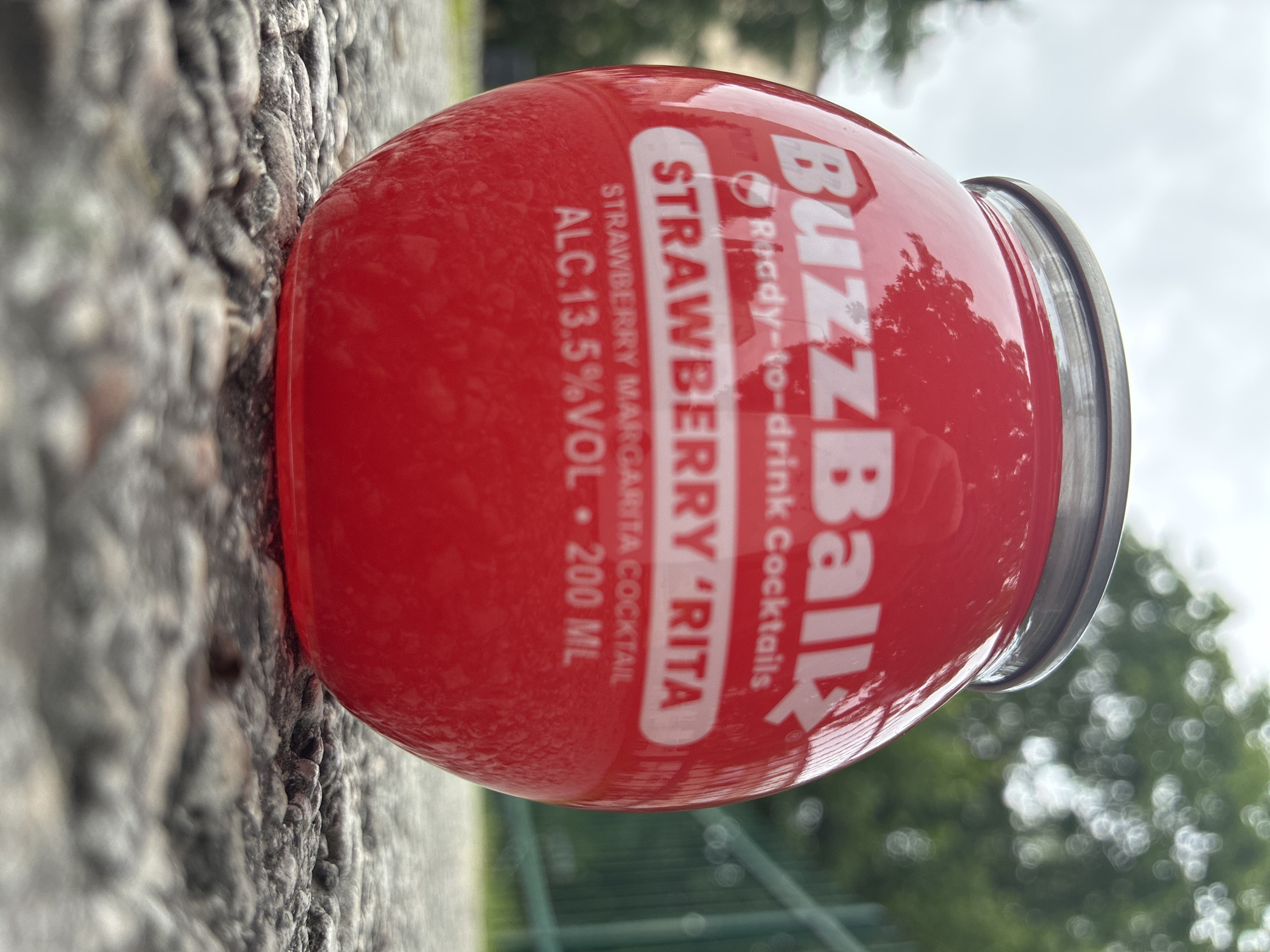 A red, spherical can of BuzzBall Strawberry 'Rita ready-to-drink cocktail sits on a gravel surface. Text on the can reads "BuzzBall," "Ready-to-drink Cocktails," "STRAWBERRY 'RITA," "STRAWBERRY MARGARITA COCKTAIL," "ALC. 13.5% VOL," and "200 ML." The background is blurred, showing green foliage and a cloudy sky. The location is not specified to be in Haapsalu, Estonia.