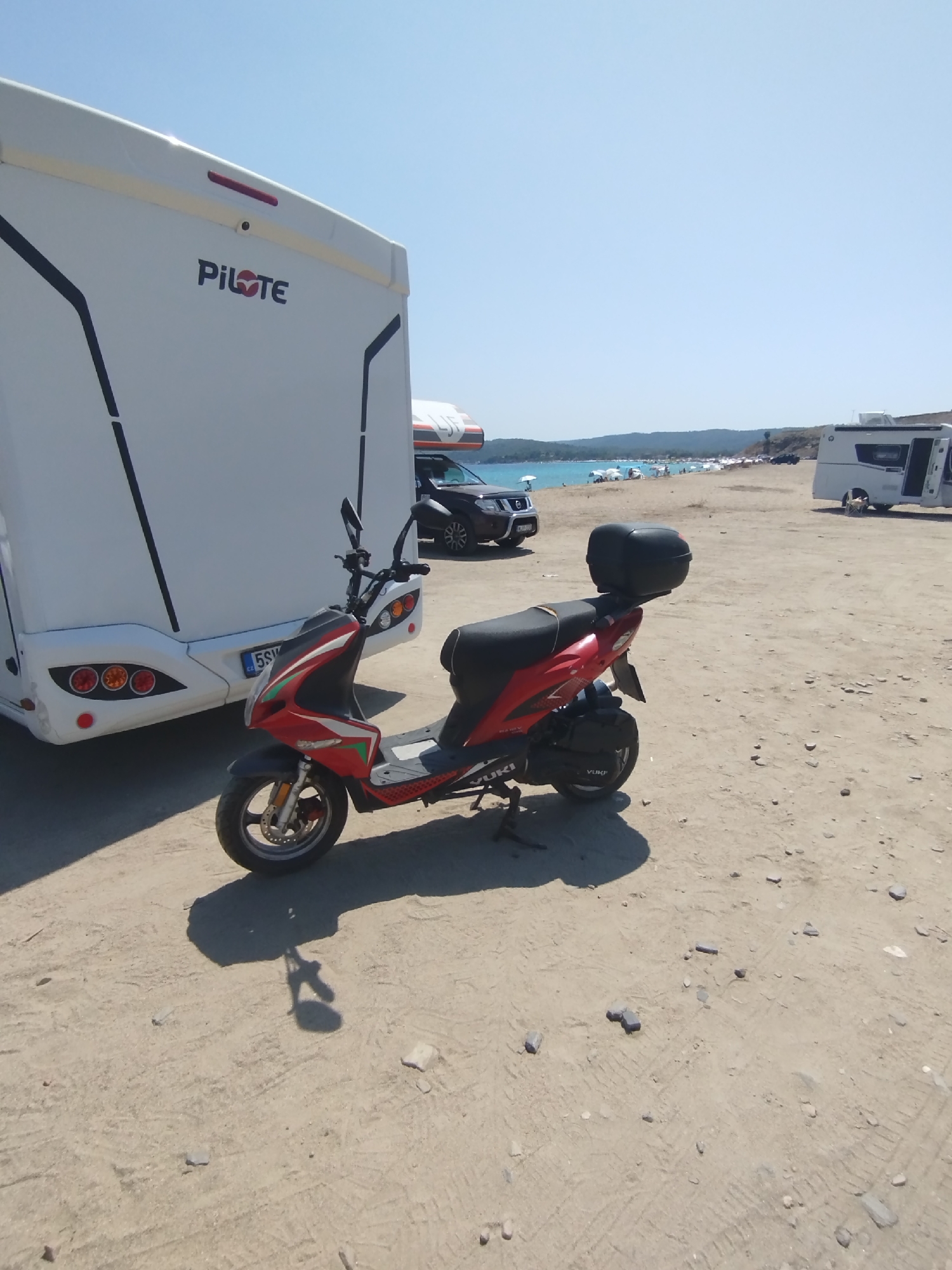This image captures a sunny day scene, likely at a beach or coastal area within the Sithonia Municipal Unit, Greece. In the foreground, a red and black scooter is parked on a sandy, gravelly surface, casting a shadow. Behind the scooter, a large white campervan with "PILOTE" branding is visible. To the right, another white caravan or camper is parked, with its door open. In the background, a body of water, likely the sea, is visible, with a beach populated by numerous umbrellas and people. Hills and vegetation form the backdrop to the coastline. The sky is clear and blue.