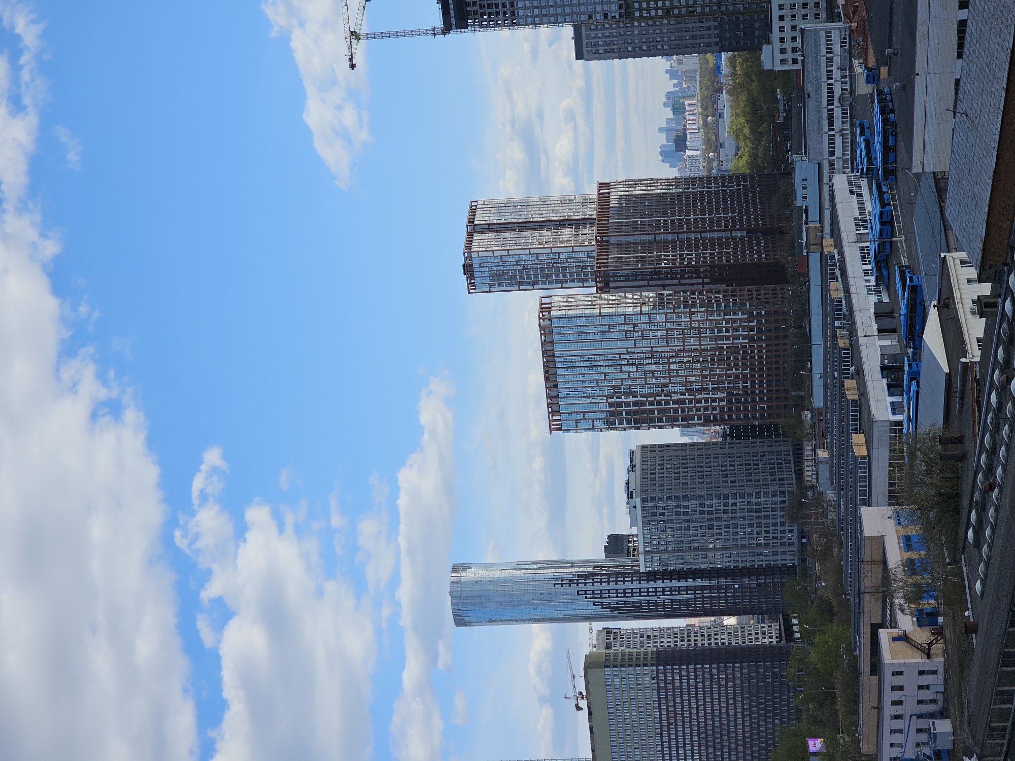 The image depicts a skyline of Moscow, Russia, viewed from a high vantage point, dominated by modern high-rise buildings and a blue sky with scattered clouds. Several skyscrapers of varying heights and designs are present, with facades of glass and metal. A construction crane is visible near one of the taller buildings. In the background, a line of buildings is visible on the horizon. The lower right section of the image shows portions of lower buildings and infrastructure.