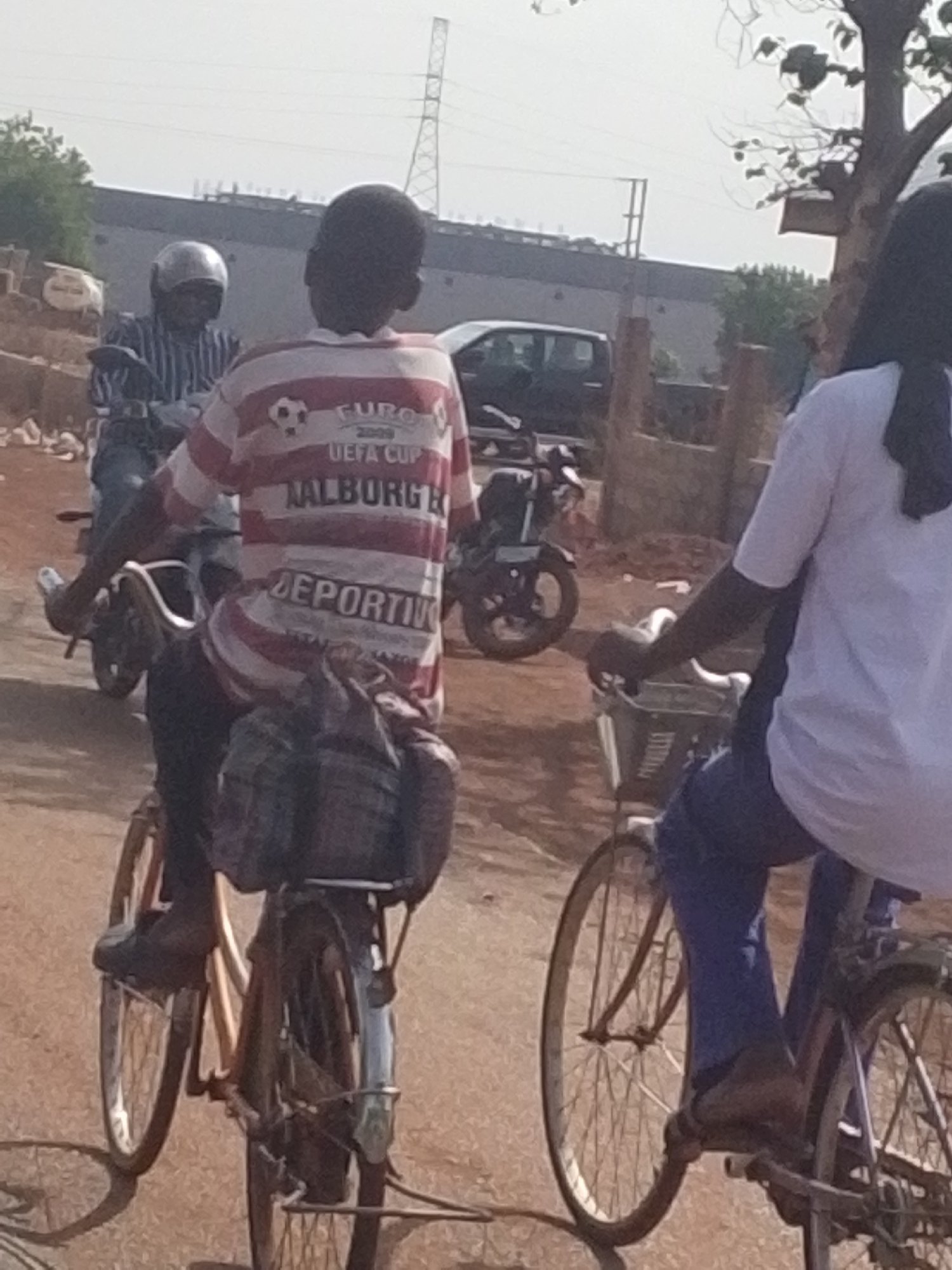 The image displays an outdoor scene with three individuals in motion, under bright daylight. In the foreground, positioned on the right, a person with dark hair, wearing a white t-shirt, blue jeans, and sandals, is riding a bicycle equipped with a front basket. Behind them, in the center, another individual is riding a bicycle, viewed from the back. This person wears a red and white striped t-shirt featuring a soccer ball logo and text that reads "EURO 2009 UEFA CUP," "AALBORG," and "DEPORTIVO." A dark bag or package is strapped to the rear of their bicycle seat. To the left and slightly behind, a third person is seen on a motorcycle, wearing a light-colored helmet and a striped shirt.
The setting appears to be an unpaved or dusty road, suggesting a semi-urban or rural environment. In the background, there are industrial-looking buildings or structures, and a tall power transmission pylon is visible against a clear, bright sky. The presence of trees on both sides indicates an open area. The activity depicted is people commuting or traveling by bicycle and motorcycle. The bright lighting suggests it is daytime.