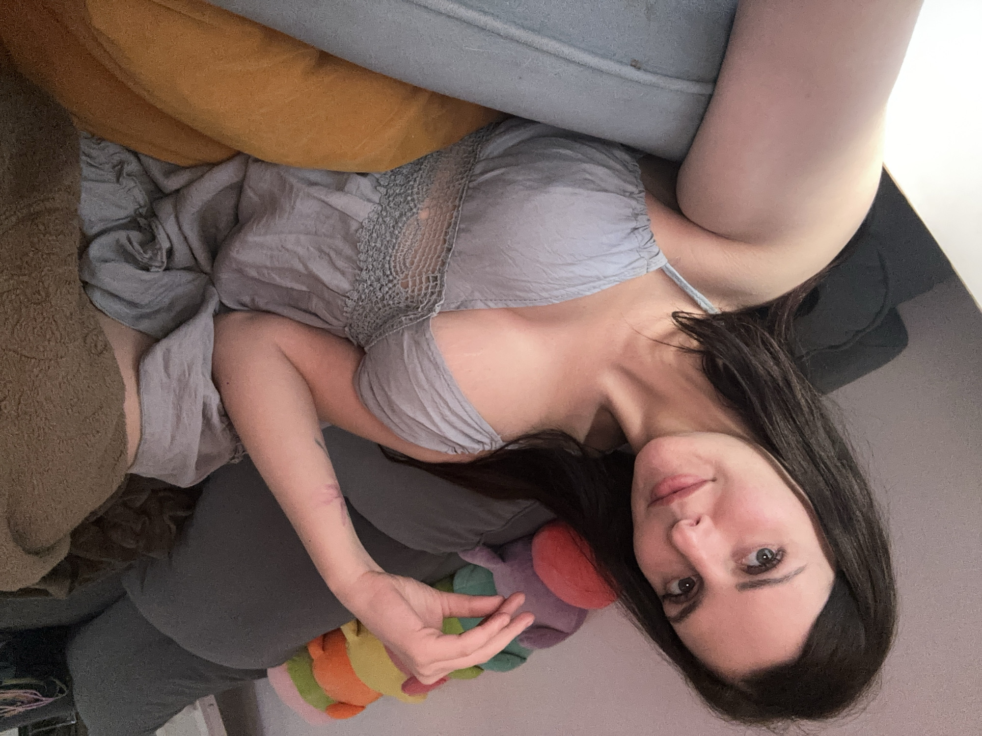 A young woman with long, dark hair is lying on a couch, angled towards the camera. She is wearing a light gray dress with delicate lace detailing around the neckline. Her right arm is bent, with her hand resting on her lap, and her left arm is extended along the couch. There is a colorful stuffed caterpillar toy visible near her lower body.
The couch appears to be covered in various textured fabrics. A mustard-yellow pillow is visible behind her head, and a light blue pillow is above that. To her left, a brown, textured object, possibly another pillow or blanket, is also visible. The background is a light, neutral color, suggesting an indoor setting. The lighting is soft, and the overall mood seems relaxed and casual. There is no visible text in the image.