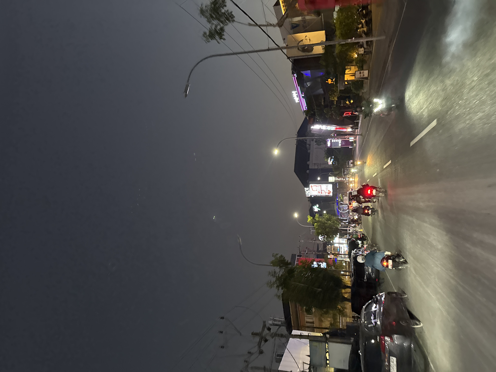 The image depicts a street scene at night in Kuta, Indonesia. The sky is a dark gray, suggesting twilight or a heavily overcast night. A road runs through the center of the frame, with multiple lanes marked by white lines. On either side of the road are buildings with illuminated signs and storefronts, indicating commercial activity. Streetlights are present, casting pools of light on the road.
Several people are on motorcycles, navigating the road. Some are riding individually, while others appear to be in small groups. The motorcycles have their headlights on, illuminating the path ahead. A few cars are parked on the left side of the road, and one car is moving in the same direction as the motorcycles. The buildings vary in height and architectural style, with some featuring modern designs and neon lighting. Trees and vegetation are visible in some areas, adding a touch of nature to the urban landscape. The overall atmosphere is that of a busy street at night, with ongoing traffic and commercial life.