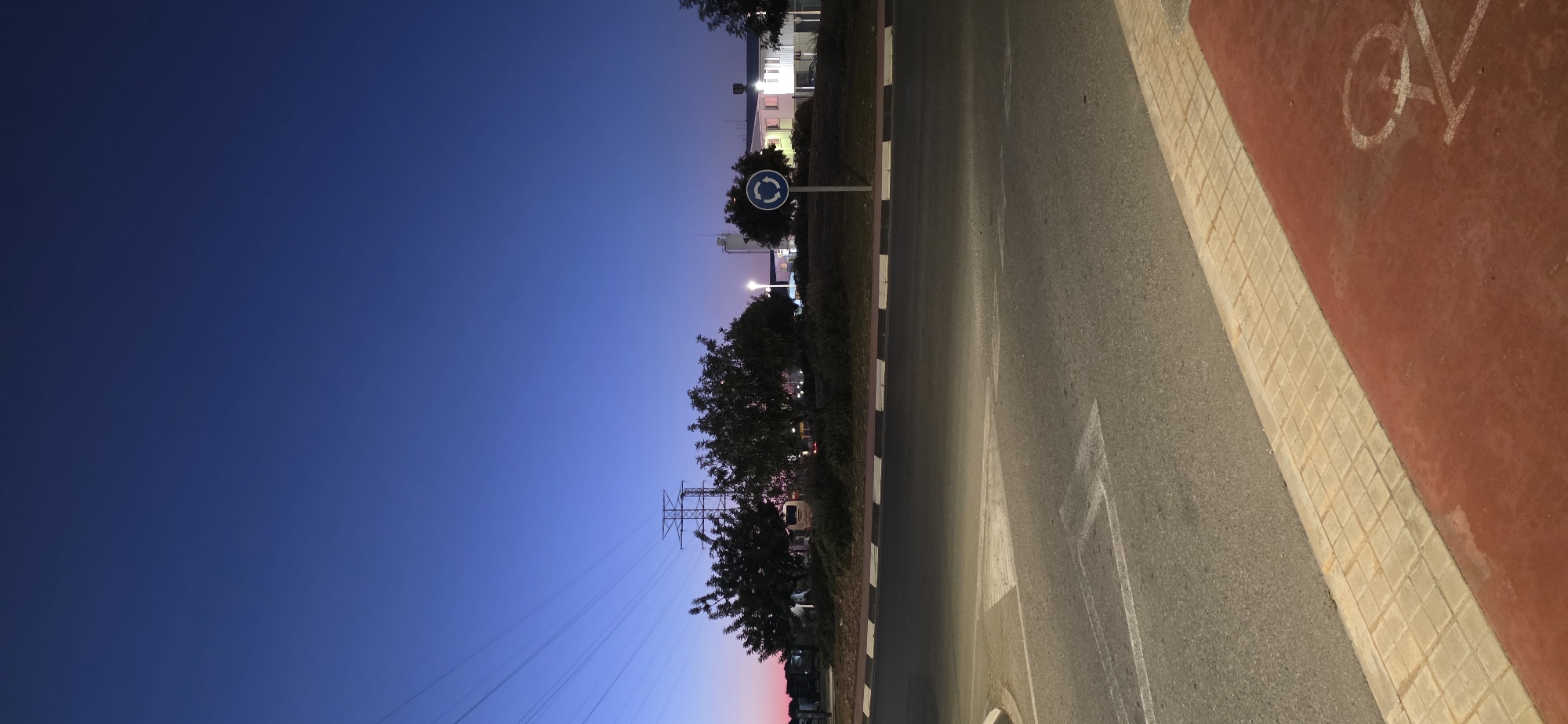 The image depicts a street in Sagunto, Spain, at dusk. The sky transitions from deep blue to a faint pink at the horizon. On the left side of the frame, trees line the road, and a roundabout sign is visible. Power lines extend into the distance. The right side of the image shows a paved road with a painted arrow and a red-colored bike lane. A tiled curb separates the road from the bike lane.