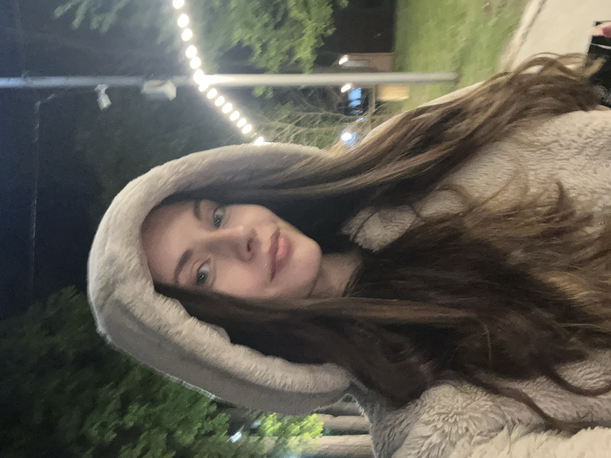 A woman with long brown hair and fair skin is wearing a light gray fuzzy hooded jacket. She is looking at the camera with a slight smile. The photo appears to be taken at night, with string lights blurred in the background. There are also trees and a pole visible in the background. The location appears to be an outdoor setting within a city.