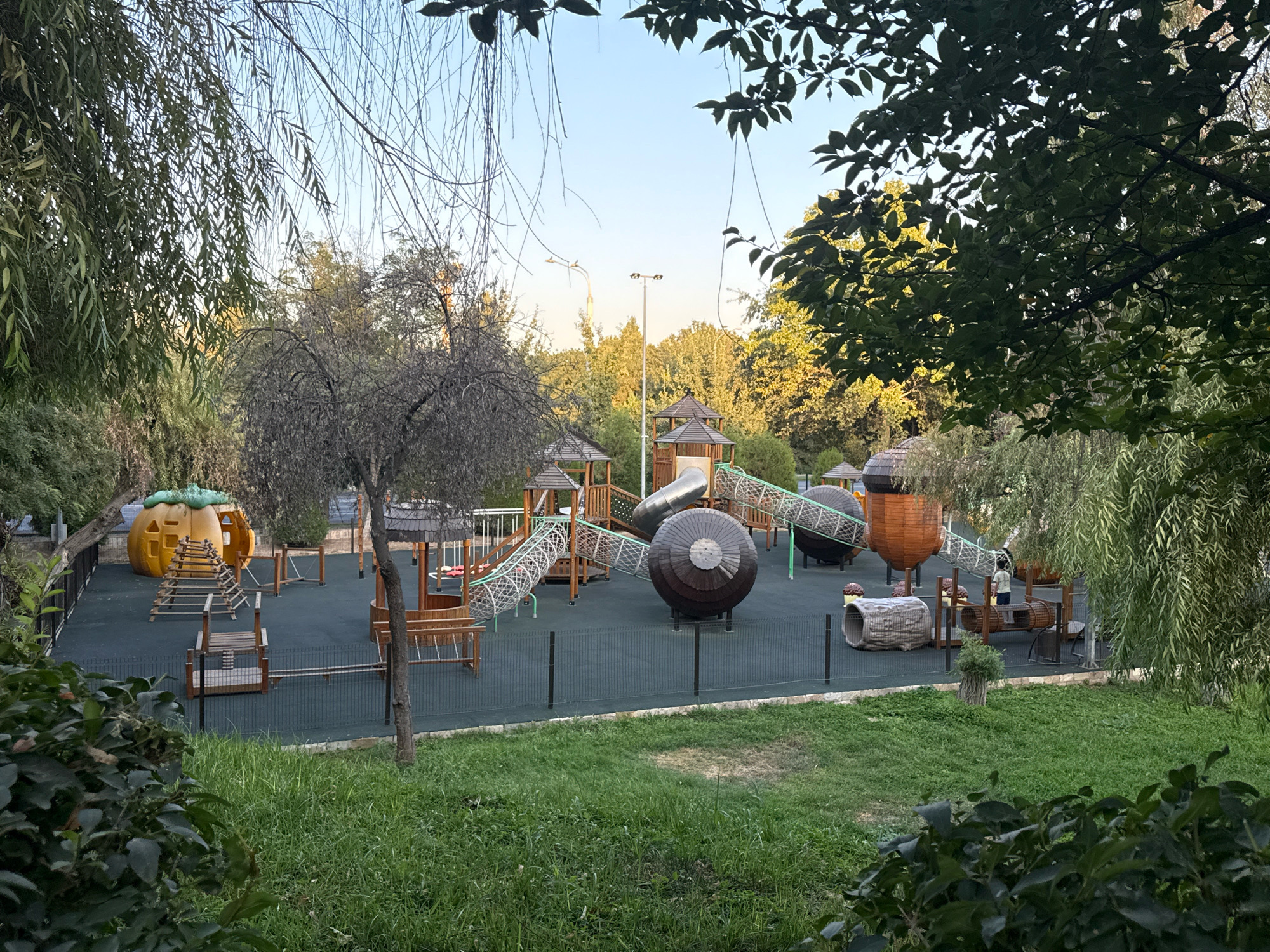 An outdoor children's playground is visible in the image, situated in Tashkent, Uzbekistan. The playground features various wooden and plastic structures, including slides, climbing frames, tunnels, and a distinctive large, orange, pumpkin-shaped playhouse. The ground surface is covered with dark green safety matting, and a black metal fence encloses the play area. Surrounding the playground is a strip of green grass, with trees and foliage framing the scene, creating a park-like atmosphere. The sky above is clear and pale blue, suggesting it is daytime, possibly late afternoon due to the lighting. There are no people clearly visible within the playground itself, though a child is partially visible near a wooden tunnel structure on the right side of the playground. The overall impression is one of a well-maintained, modern playground set within a natural, leafy environment.