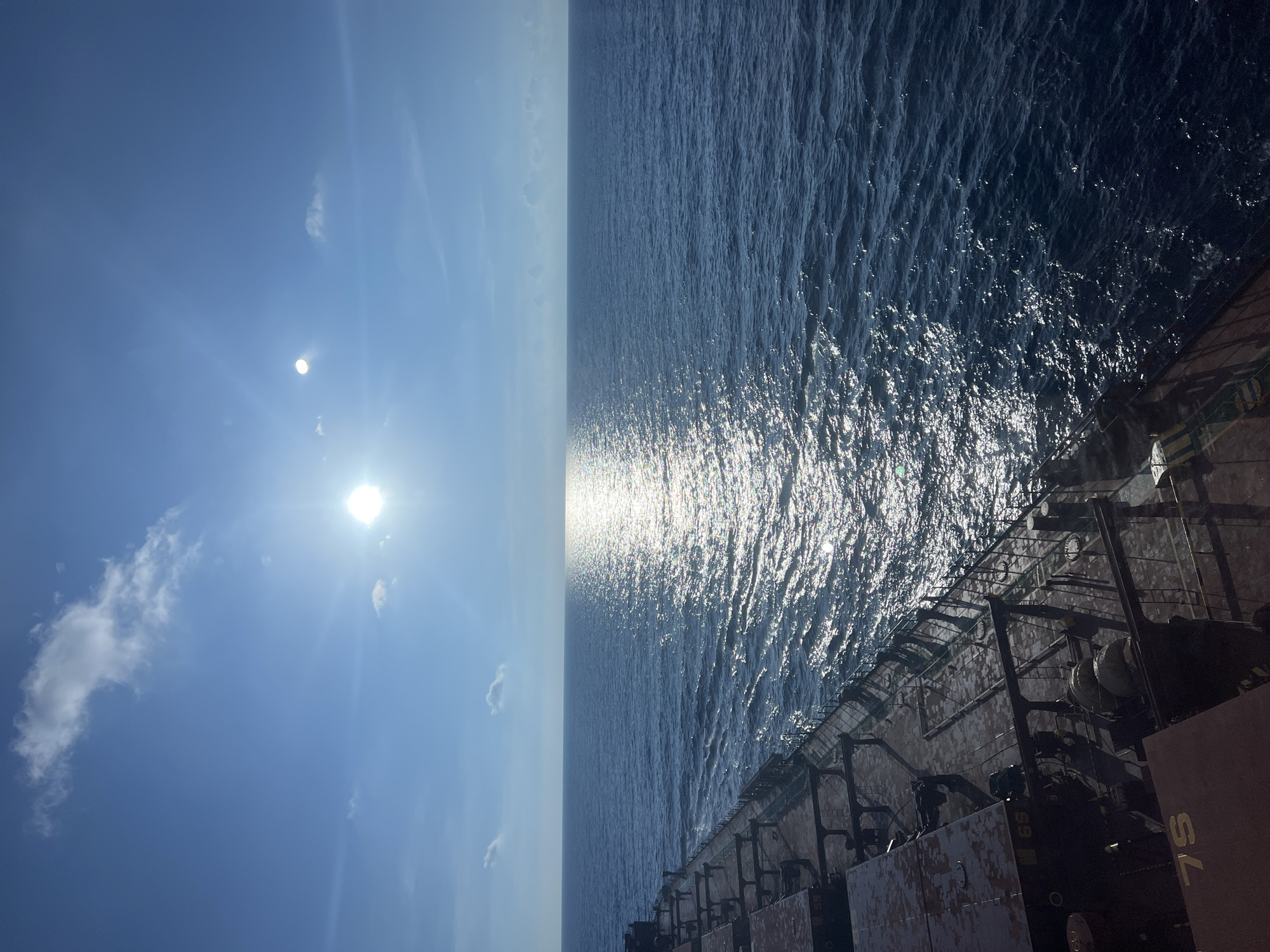 An eye-level, wide-angle shot captures the vast expanse of the ocean under a bright, sunny sky. The sun, positioned in the upper left quadrant, casts a brilliant, shimmering reflection across the deep blue water. A few scattered clouds dot the sky. In the foreground, the deck of a large ship, likely a cargo vessel, is visible, extending from the bottom left corner towards the right. The deck appears weathered, with visible markings and railings. The overall scene conveys a sense of openness and travel at sea.