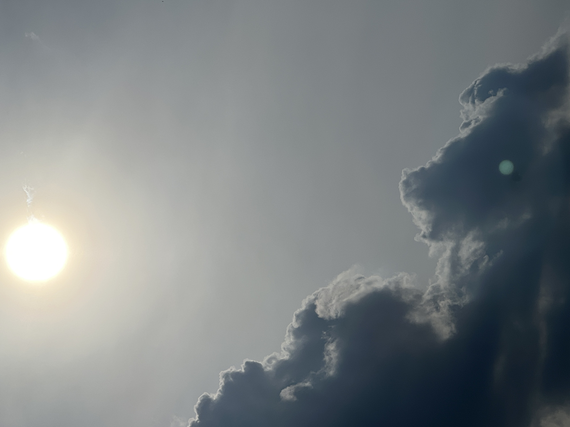 A bright, hazy sun is visible in the upper left quadrant of the image. Below the sun, dark, dramatic clouds fill the lower half of the frame, with lighter clouds closer to the sun. The sky is overcast and gray. The scene is not specific to any location within Warsaw, Poland, or any city, as it depicts only the sky and clouds. There are no people, subjects, objects, or actions depicted.