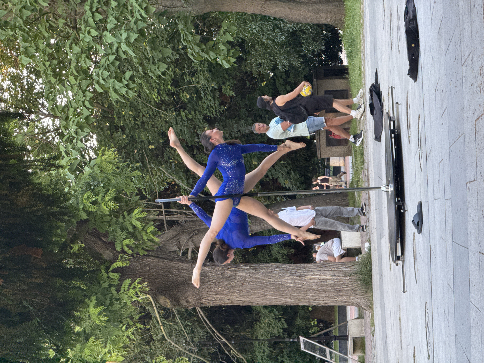 Two women, appearing to be identical, perform a synchronized aerial routine on a portable pole in a public park in Varna, Bulgaria. They wear matching royal blue, long-sleeved bodysuits with reflective embellishments and are barefoot. Their bodies are held horizontally, approximately parallel to the ground, with legs extended in opposite directions and arms securing them to the silver-colored vertical pole. One woman faces left, the other right. The pole is affixed to a circular black base system with stabilizing bars extending onto the light grey paved surface. A pair of black flip-flops rests on the pavement near the pole's base, and two black equipment bags lie further to the right.
In the background, a large, mature tree with rough bark stands prominently behind the performers. Other dense green foliage from various trees and bushes forms the backdrop, suggesting a well-vegetated park environment. The ground consists of light-colored paving stones.
Several individuals are visible in the mid-ground and background. To the left, a man in a light t-shirt and shorts sits on a light-colored stone bench. Centered behind the pole, a man in a light t-shirt and denim shorts stands with arms crossed, observing. To his right, another man, wearing a black t-shirt, black shorts, and a black baseball cap, walks while carrying a yellow and black ball and a black backpack. Further right and in the distance, two women walk away from the scene. An A-frame sign with a white panel is visible on the far left. The scene is illuminated by natural light, suggesting daytime.