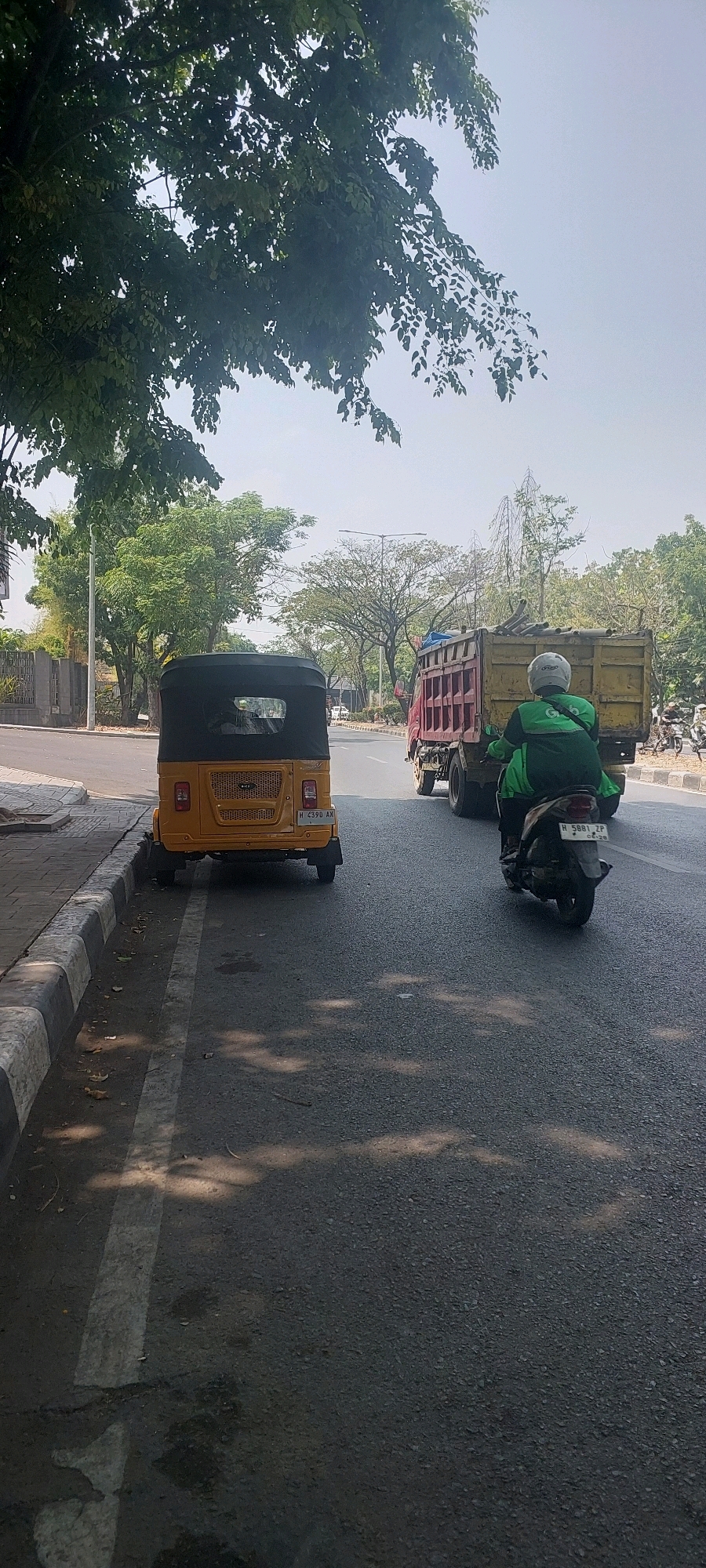 The image depicts a street scene in Semarang, Indonesia, on a bright, sunny day. The foreground shows the rear of a yellow auto-rickshaw parked along the side of a paved road. The auto-rickshaw has a black canvas roof and a visible license plate. To the right of the auto-rickshaw, a motorcycle carrying a person in a green jacket with a white helmet is moving along the road. The rider's jacket features a prominent "Grab" logo, indicating a ride-hailing service. Further down the road, a large red and yellow truck is in motion, appearing to be loaded with materials. The surrounding environment includes a sidewalk with a distinctive black and white border, trees with lush green foliage lining the road, and buildings in the background. The sky is a clear, pale blue with a few wispy clouds, and sunlight casts shadows on the road surface. The overall impression is of a typical urban street during the daytime in Indonesia.