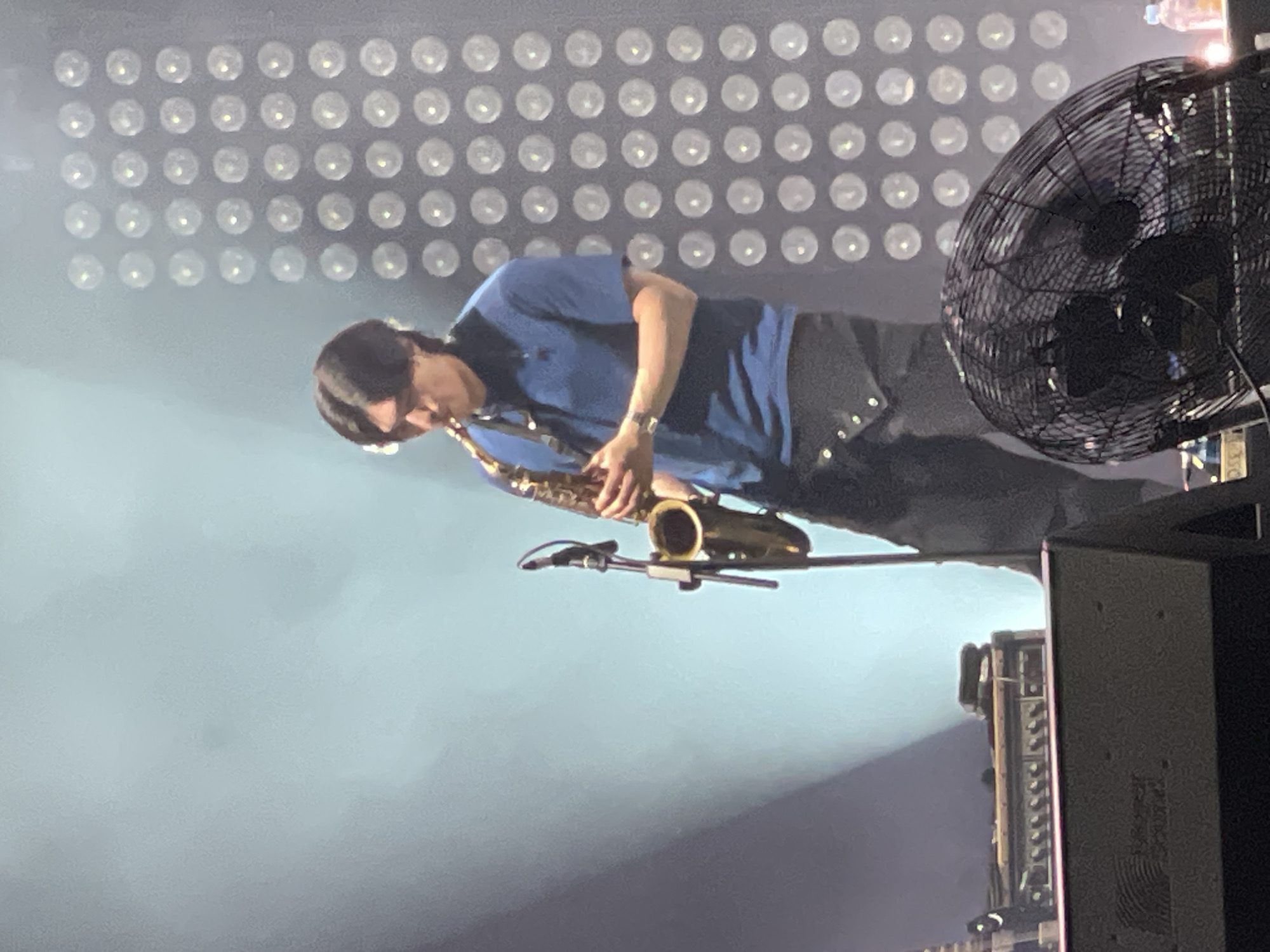 A musician plays a saxophone on a stage. He is wearing a blue short-sleeved shirt and dark pants. A microphone is positioned in front of the instrument. Behind him is a grid of illuminated lights. To his right is a large fan and stage equipment. The location is Salembaran, Indonesia.