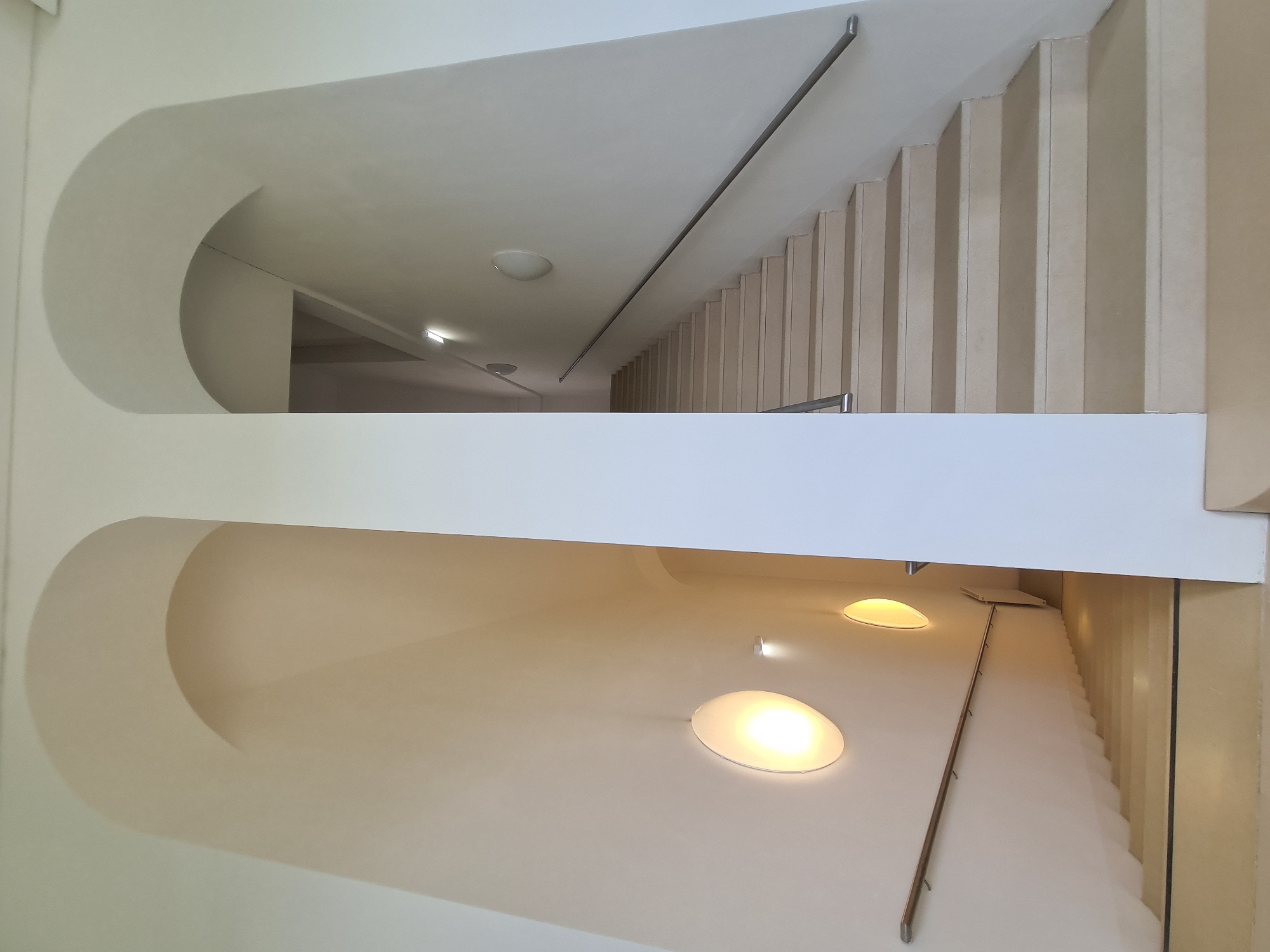 This is a vertical, eye-level, full shot of an interior staircase. The staircase is a grand, curving design with two distinct ascending paths. The left path is illuminated by two circular wall sconces, one of which is brightly lit. The right path is more dimly lit, with only a hint of light from further up the stairs and a wall-mounted light fixture visible. Both staircases feature pale, wide steps and are lined with handrails. The walls are a smooth, off-white color and feature large arched openings. The environment appears to be clean and modern. There are no people visible in the image. The scene does not provide any specific details to indicate the location in Vienna, Austria.
