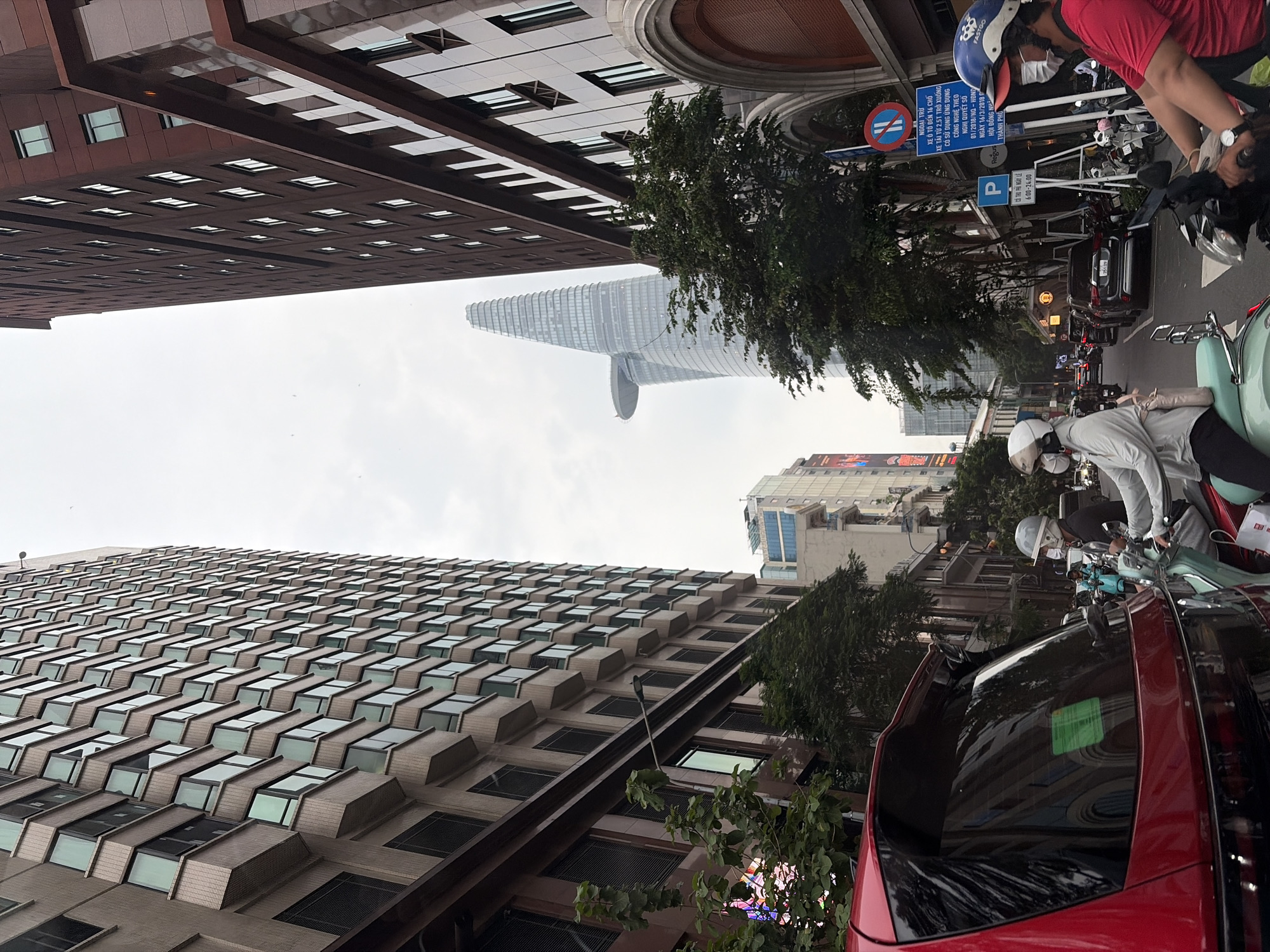 The image depicts a street scene in the city of Thủ Đức, Vietnam, on an overcast day. Tall buildings line both sides of the street, with a prominent skyscraper featuring a helipad visible in the distance. A red car is parked in the foreground on the left, with its rear visible. Several people are on motorbikes, some wearing helmets and face masks, navigating the traffic. Traffic signs are visible on the right side of the street, including parking and traffic regulation signs. Trees and streetlights are also present along the urban thoroughfare.