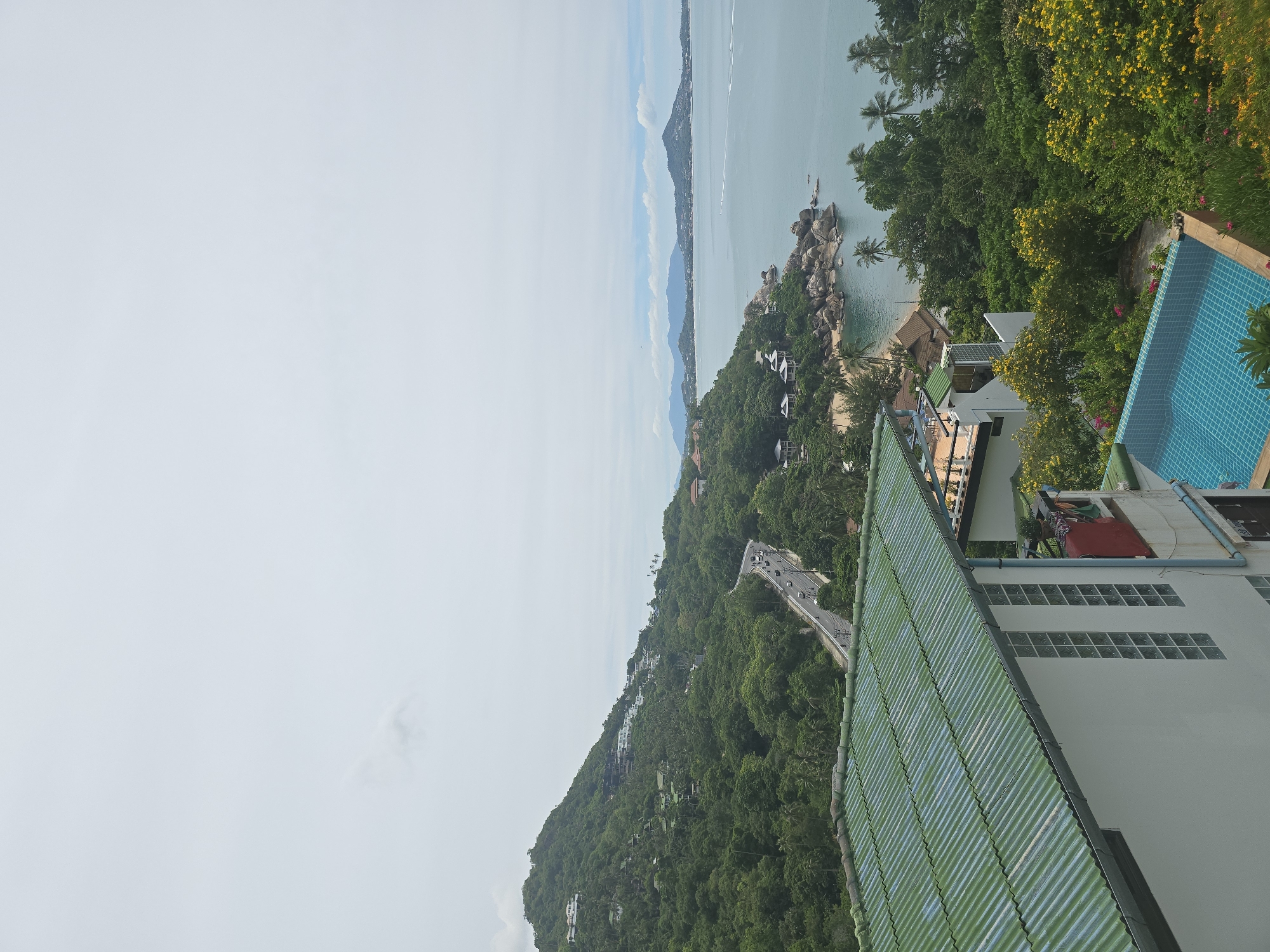 A high-angle, wide shot captures a coastal landscape with a prominent green-roofed building in the foreground. The building has white walls and a blue-tiled swimming pool. Beyond the building, a lush, green hillside is covered in trees and dotted with white houses. A winding road with cars is visible on the hillside. The ocean stretches to the horizon, with distant islands and a cloudy sky overhead. The scene appears to be a resort or residential area in a tropical location.
