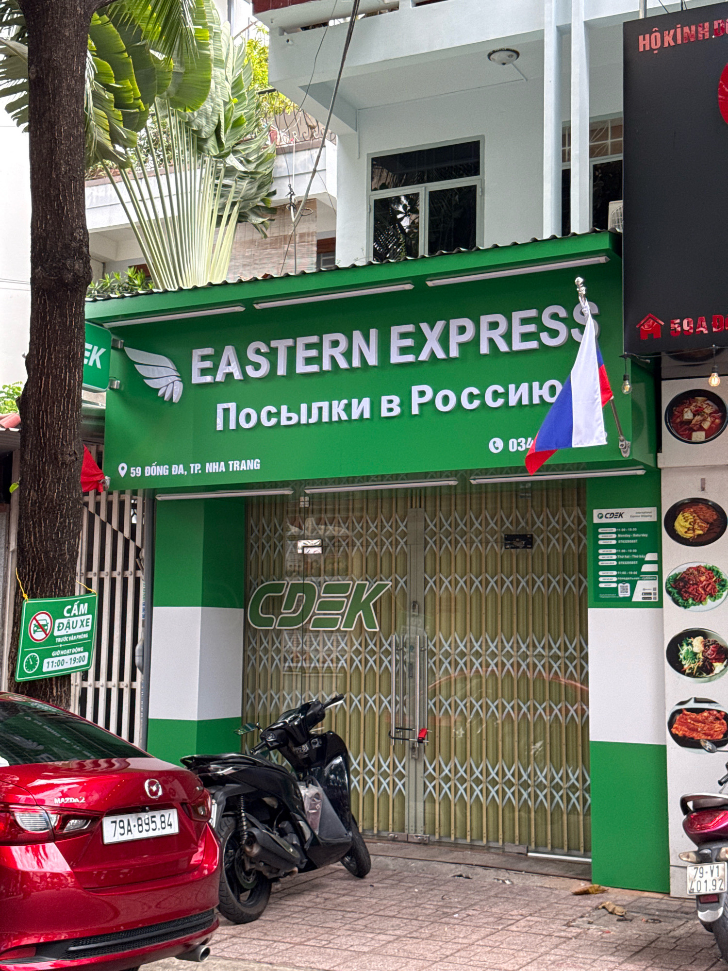 The image captures the exterior of an Eastern Express logistics company branch in Nha Trang, Vietnam. The green storefront features the company logo and text in English and Russian, along with the address "59 ĐỒNG ĐA, TP. NHA TRANG." A Russian flag is displayed on a pole in front of the building. To the left, a green sign indicates "CẤM ĐẬU XE" (No Parking) with operating hours listed as 11:00-19:00. In the foreground, a red Mazda 2 car is parked, and a black motorcycle is positioned near the entrance. The storefront itself is a rolling metal gate with a "CDEK" logo visible. A vertical stack of circular images depicting Asian cuisine is mounted on the right side of the building. The overall scene is a street-level view of a business establishment in a city environment.