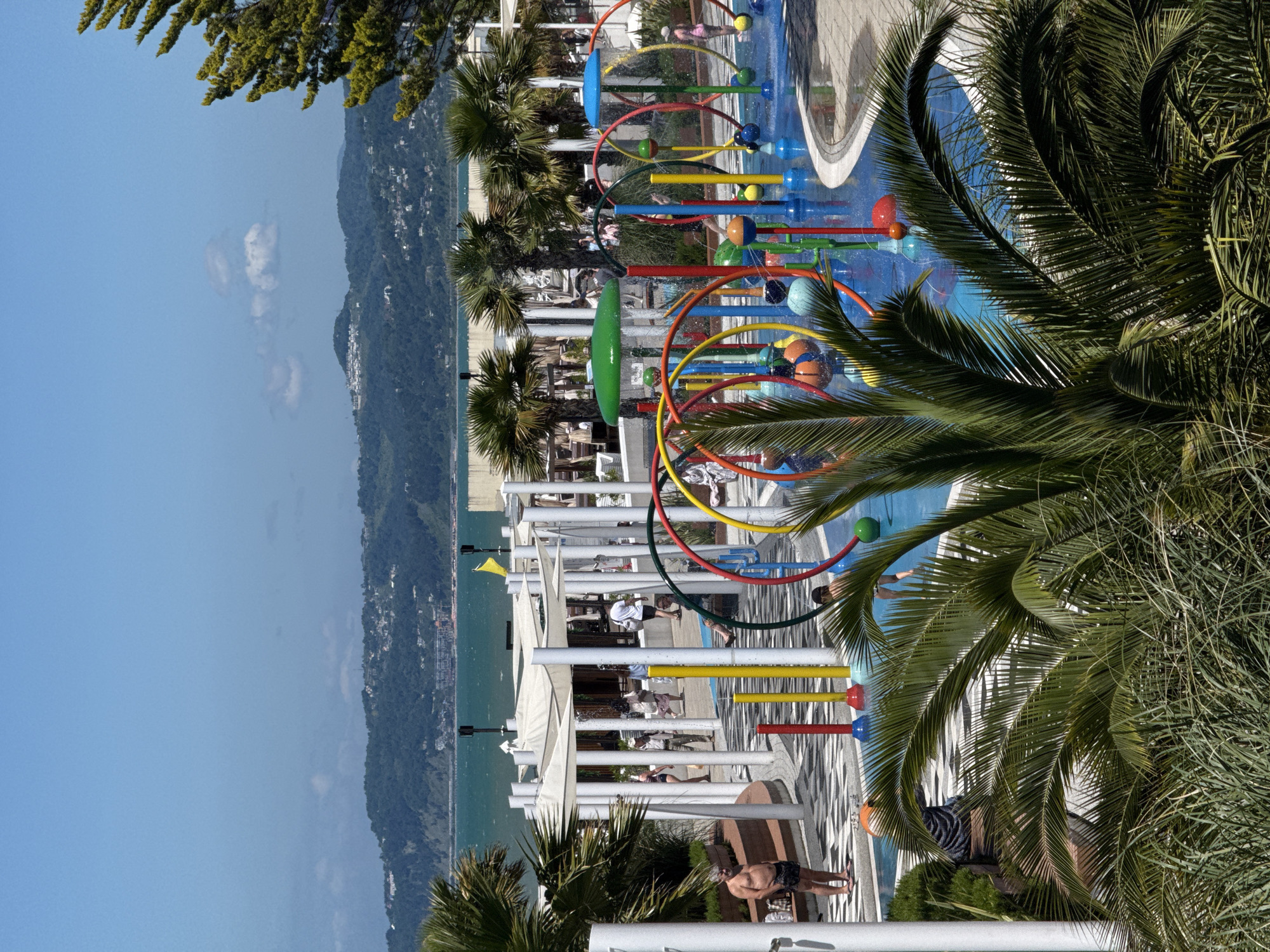 This image depicts a vibrant, sun-drenched coastal resort scene, likely in Sochi, Russia. The perspective is slightly elevated, looking down over a multi-colored water play area towards the sea and mountains.
In the foreground and mid-ground, a lively water park features bright blue water, adorned with an array of colorful elements including red, yellow, and blue rings, spheres, and spray features. A prominent green, arch-like structure is also part of the play equipment. Several lush green palm trees frame the scene, their fronds extending into the shot, casting some dappled shade. People, including adults and children, are visible enjoying the water features or relaxing nearby. One man in dark swim trunks stands casually near white support pillars in the lower mid-ground.
Beyond the water park, a series of white sunshade structures and loungers indicate an outdoor dining or relaxation area, with more individuals visible under the shade. The backdrop features a expansive, calm turquoise sea. The distant horizon is lined with verdant, undulating mountains covered in dense foliage, with scattered buildings and settlements dotting the slopes. The sky above is a clear, bright blue with a few soft, white clouds.
The scene conveys a sense of leisure and fun, typical of a holiday destination. The bright sunlight and clear skies suggest pleasant, warm weather during daytime. No discernible text is visible within the image.