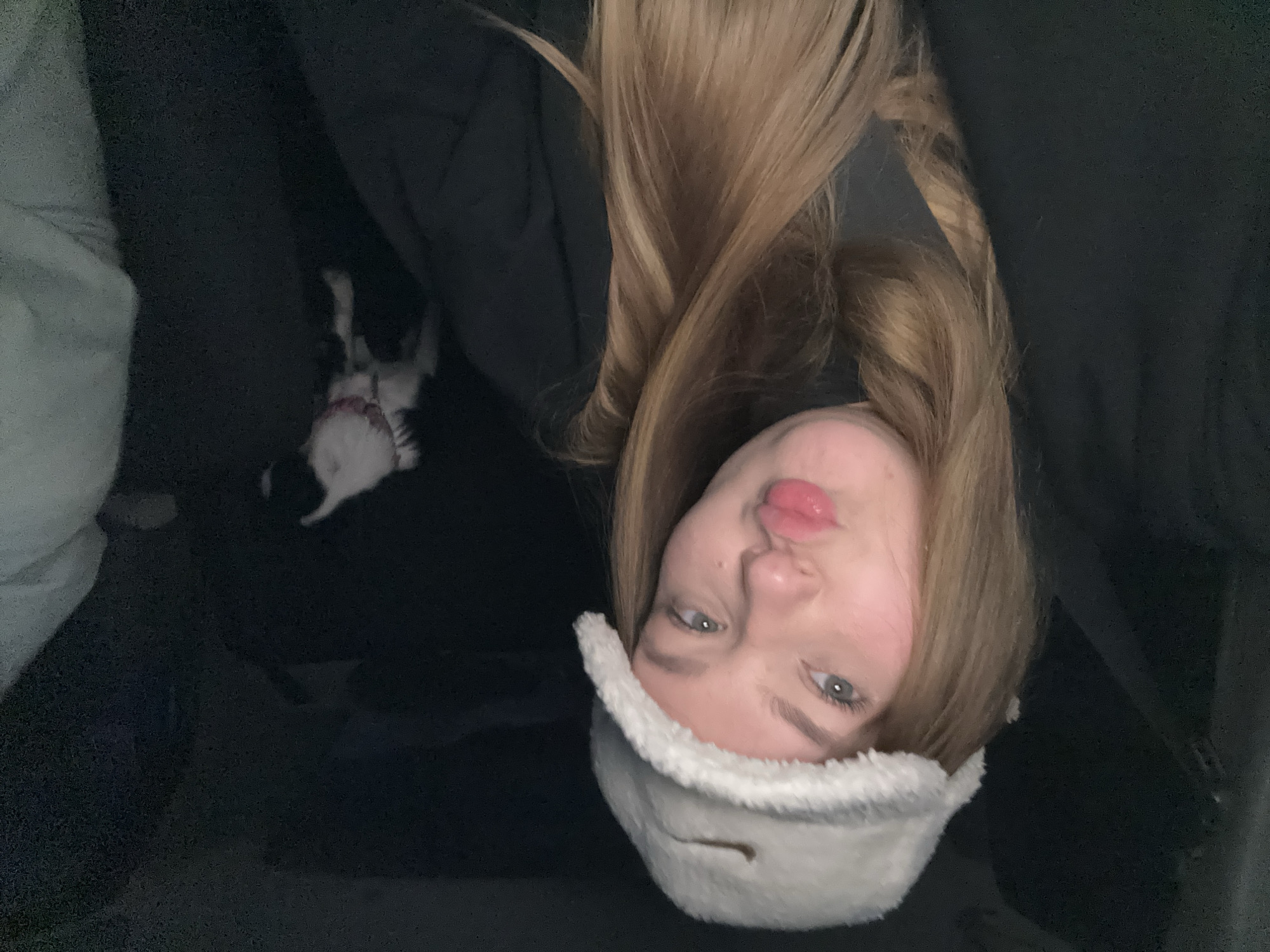 A young woman with long blonde hair is pictured indoors, wearing a white fuzzy hat. She appears to be looking at the camera with pursed lips. A small black and white dog is visible in the background, lying down. The setting appears to be a dark room, possibly a living room, with a dark-colored couch. It is not possible to know the location in the city of Chaikovsky, Russia from the image.