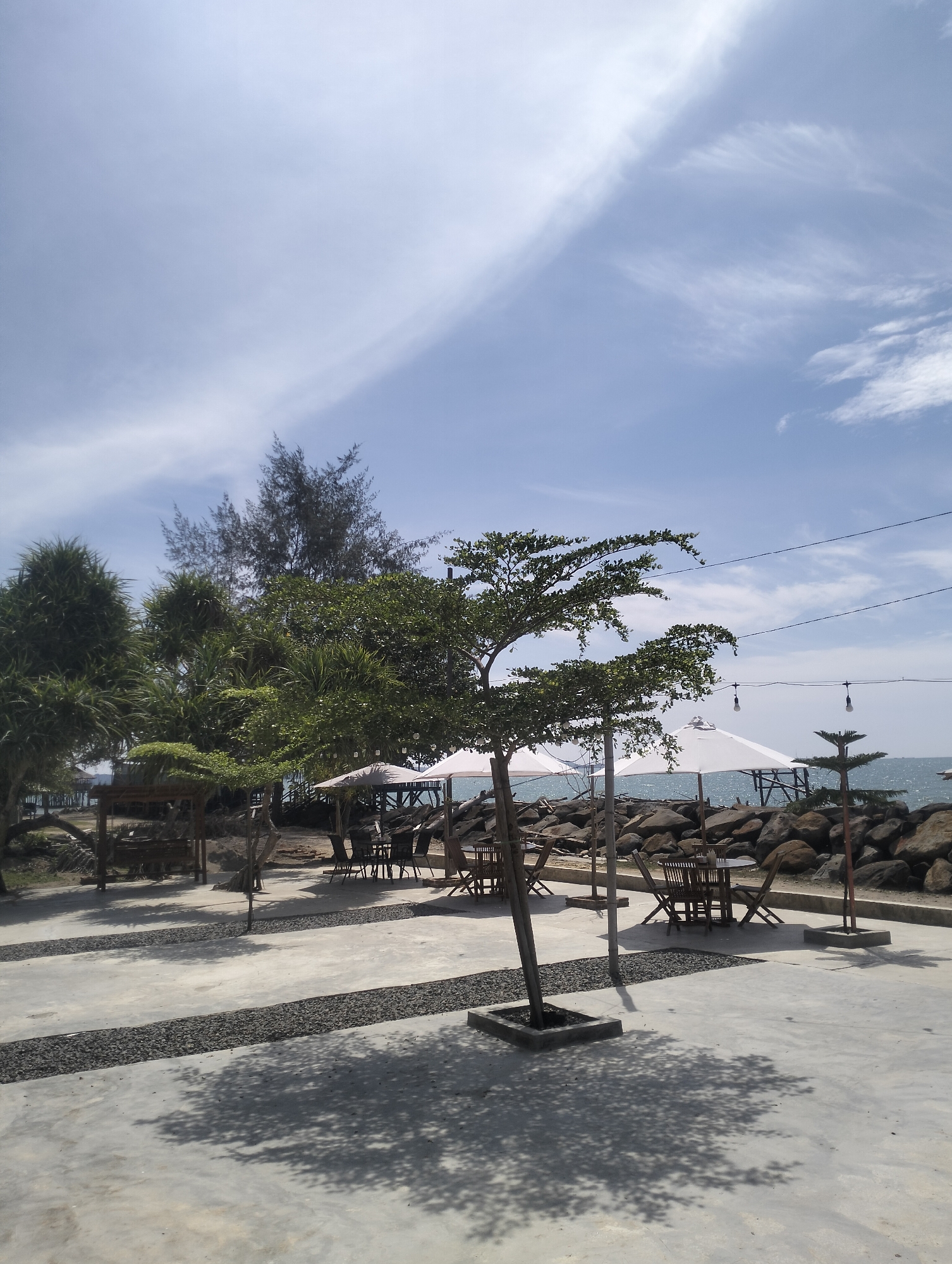 This image depicts an outdoor seating area at a seaside location, likely a cafe or restaurant, in Lancok, Indonesia. The scene is bathed in bright daylight, suggesting it is midday. The sky is a clear blue with sparse white clouds, indicating fair weather.
The foreground is dominated by a concrete patio with dark gravel pathways. Several trees are planted in square concrete bases, casting dappled shadows from the sunlight onto the patio. To the left, a wooden swing set is visible under a canopy. Further back, tables and chairs are arranged under large white umbrellas, some occupied by empty seating. These tables are made of dark wood.
Beyond the seating area, a rocky shoreline separates the patio from the sea. The ocean is calm and appears to stretch out to the horizon. In the distance, more trees, including some tall, slender ones and broader leafy trees, are scattered along the coast. There are also structures visible in the distance, possibly piers or buildings. String lights with bare bulbs hang across the scene, suggesting the area is also used in the evening. The overall atmosphere appears peaceful and relaxed, suitable for enjoying the coastal environment. No people are clearly visible, and no text is present in the image.