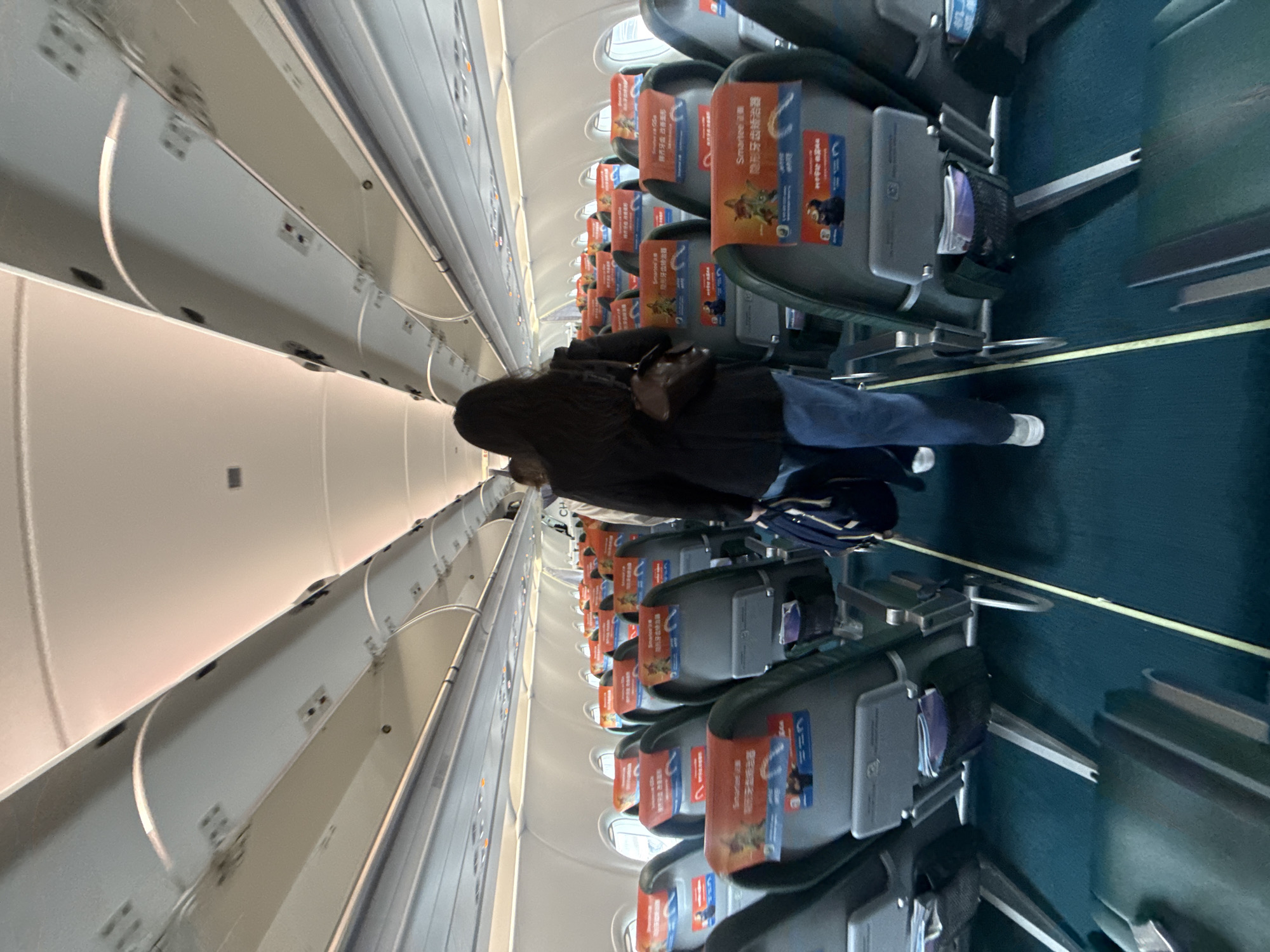 This image depicts the interior of a commercial airplane cabin, likely within the vicinity of Pudong, China. The perspective is from the aisle, looking towards the front of the plane. Several rows of empty passenger seats are visible on both sides, upholstered in a dark fabric with orange headrests displaying some form of advertising or safety information. The aisle itself is covered in a teal carpet, with a yellow line demarcating the walking path.
In the foreground, a woman with long dark hair is walking away from the camera down the aisle. She is wearing a dark jacket, blue jeans, and white sneakers. She carries a brown handbag over her shoulder and a dark blue duffel bag in her right hand. Her posture suggests she is boarding or moving through the cabin.
The ceiling of the plane features overhead compartments and lighting fixtures, consistent with a standard passenger aircraft. The windows, visible on the right side, are oval-shaped and appear to be illuminated by external light. The overall impression is one of an orderly and functional airplane environment, ready for or in the process of passenger transit.