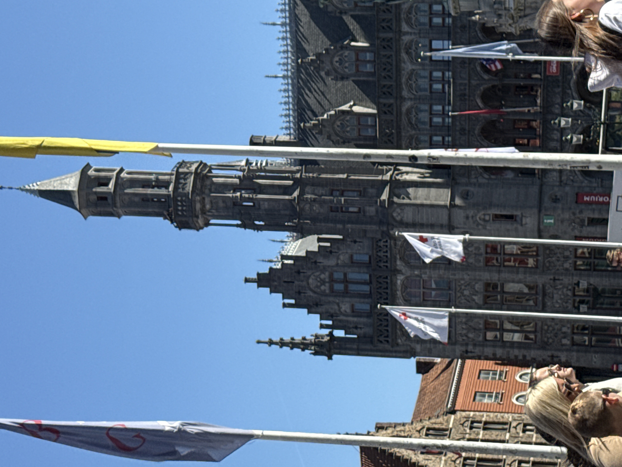 The image captures a bright, sunny day in a historic European city, identified as Bruges, Belgium, with clear blue skies overhead. The primary focus is a magnificent Gothic-style building, likely a town hall or belfry, featuring elaborate stone facades, arched windows, and a tall, slender tower with a pointed spire on the left side. The building's dark, steeply pitched roof is adorned with numerous spires and decorative elements.
In the foreground, several people are visible, mostly looking upwards. On the right side of the frame, a woman with long dark hair and a gold earring is seen gazing up. Towards the bottom right, a blonde woman with sunglasses resting on her head and a man beside her are also looking skyward, suggesting they are observing the architecture or an event. Another brick building with a red tile roof is partially visible behind the blonde woman.
Multiple banners and flags are strung horizontally across the scene. A bright yellow banner stretches across the top left. Several white flags, each bearing a prominent red cross symbol (suggesting the Red Cross), hang from poles in front of the main building. One flag appears to show part of an American flag pattern, and a grey banner with red heart-like designs is visible at the bottom left.
On a red sign attached to the grand building, the text "ORIUM" is partially visible. The scene evokes a lively tourist or public gathering spot, with visitors enjoying the architectural beauty under pleasant weather conditions.