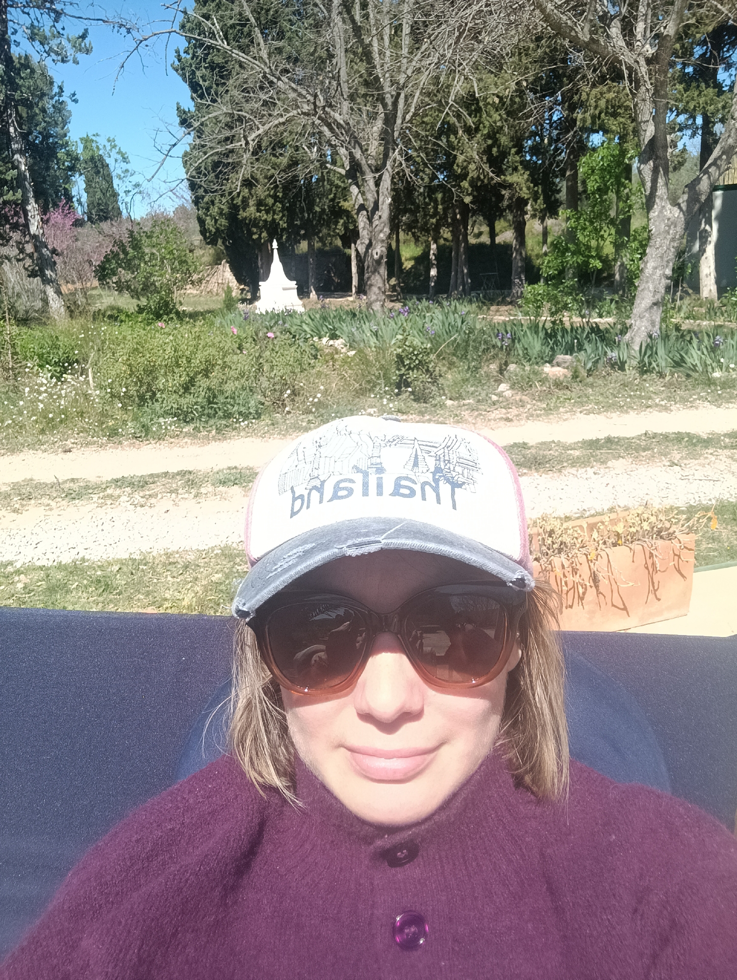 A woman wearing sunglasses and a cap with the word "basilica" (possibly Basilica de la Virgen de los Desamparados) sits outdoors. She is wearing a purple sweater. The background includes trees, grass, and a path. A small white structure is visible further back. The location is likely Jérica, Spain.