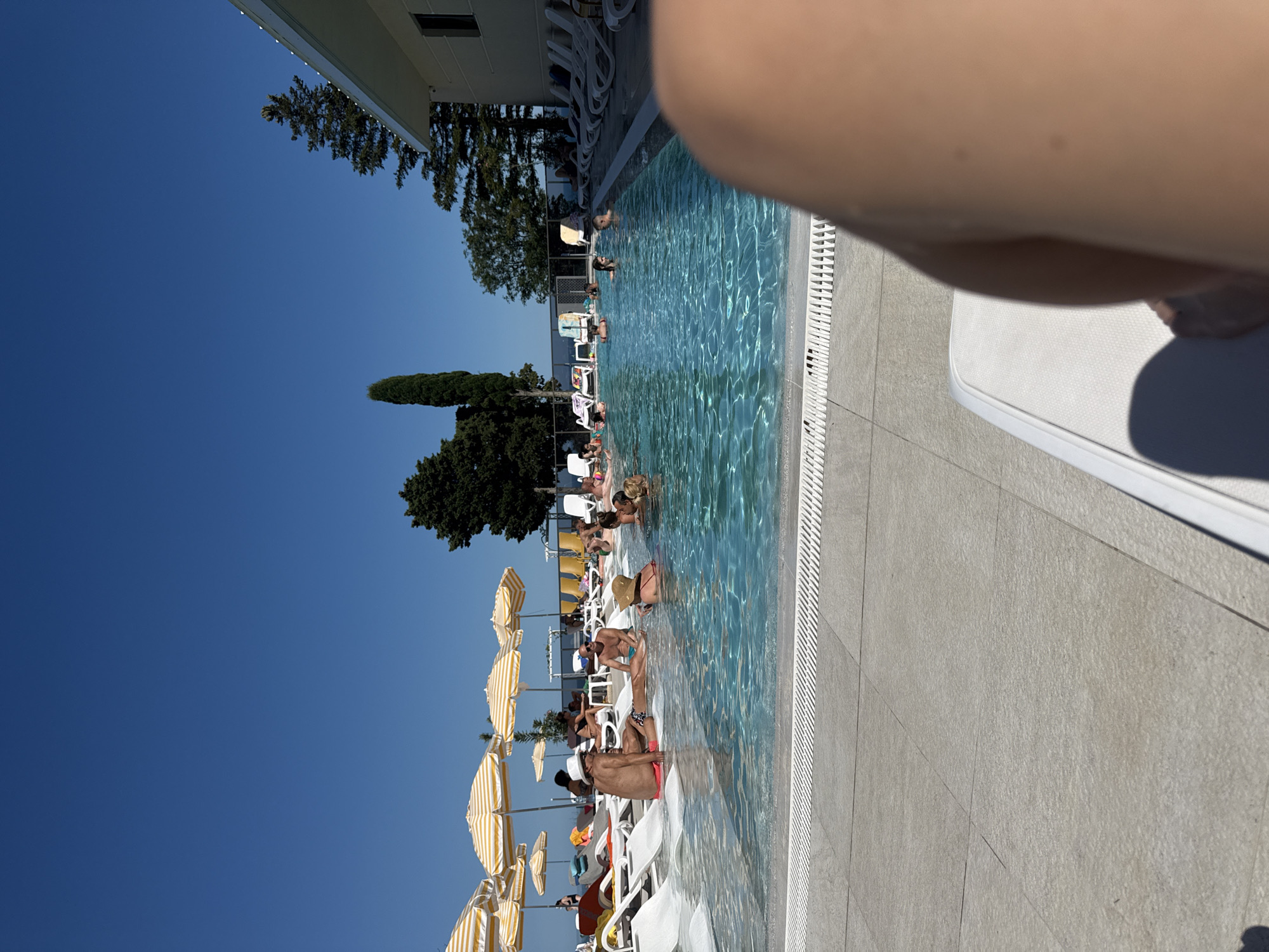 An outdoor swimming pool area on a sunny day features several people relaxing. In the foreground, a person's leg is visible, partially obscuring a white lounge chair. In the middle ground, the shallow end of the pool has several people wading and sitting. Further back, the main pool area is occupied by more individuals swimming and socializing. Lounge chairs with yellow and white striped umbrellas are set up around the pool. The background shows trees, a building with a light-colored facade, and a clear blue sky.