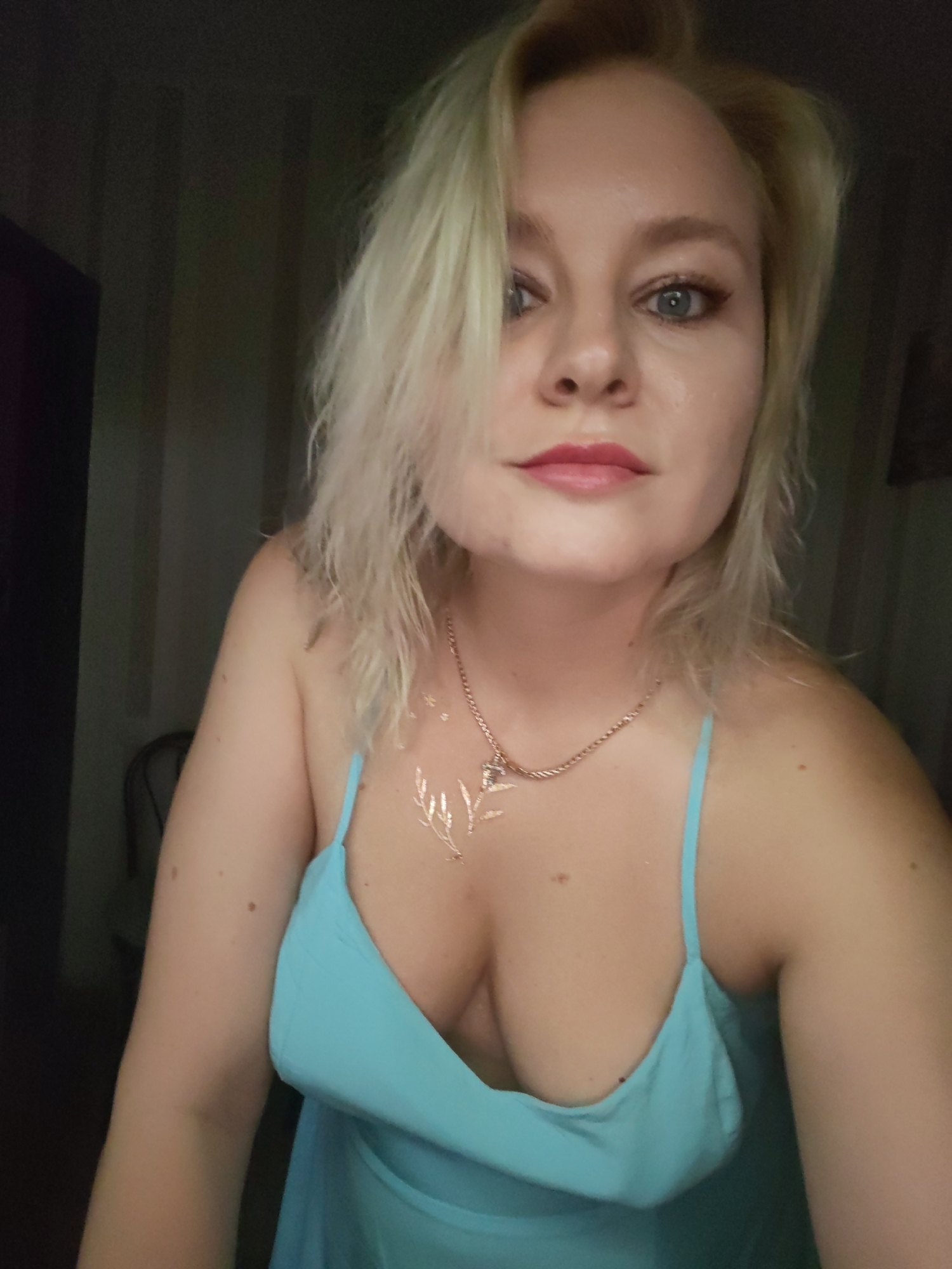 A medium shot portrait of a woman with blonde hair, wearing a turquoise dress and a necklace. The woman is looking directly at the camera. Her hair is messy, and she has light makeup on. She has a tattoo of a leafy branch on her chest. The background is dark and out of focus, with vertical stripes on the wall. The image appears to be taken indoors, in a dimly lit room. There is no specific indication of the location being Izhevsk, Russia.