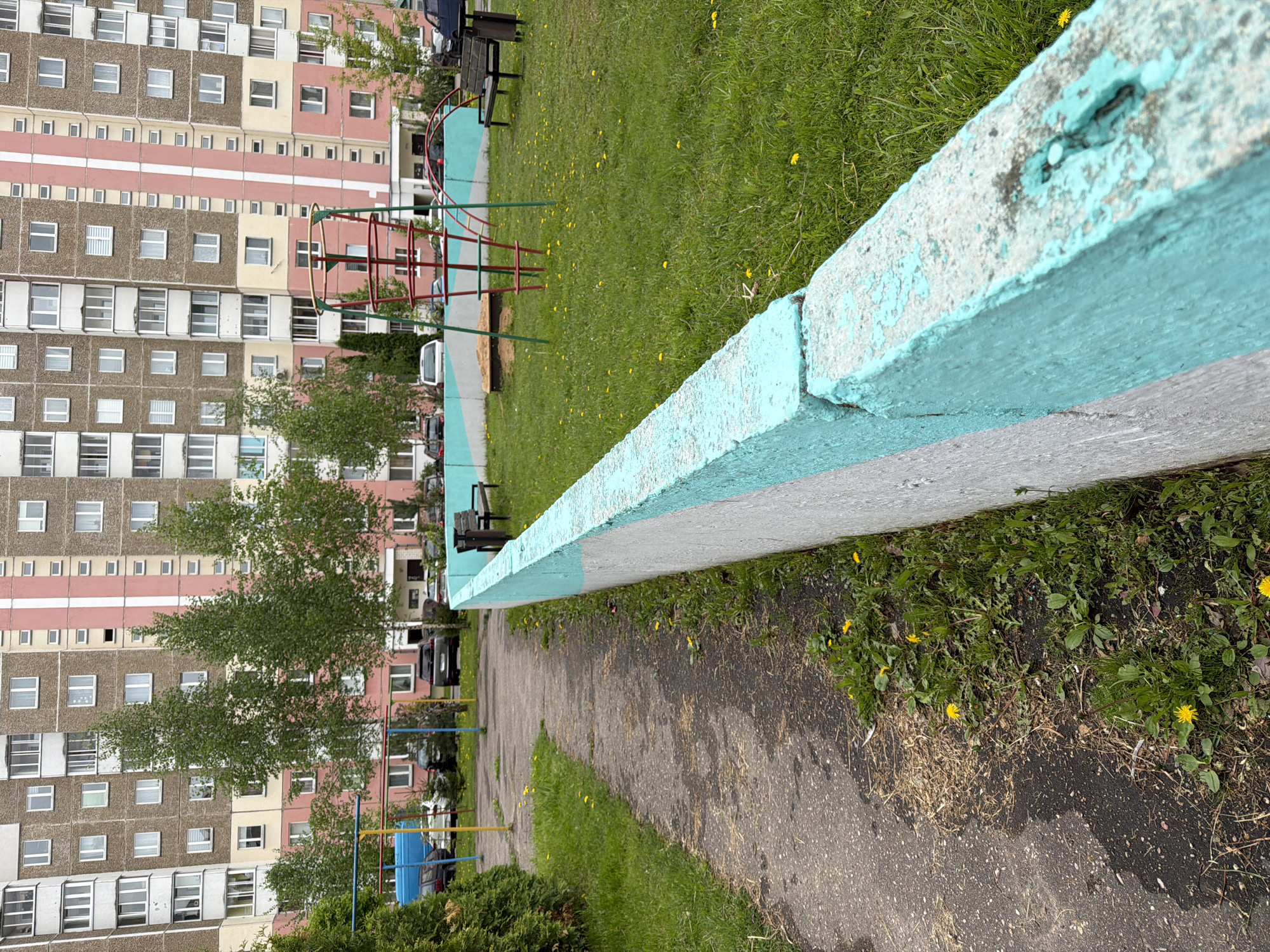 The image depicts a pathway alongside a grassy area in an urban setting, possibly in Unknown, Unknown. A low concrete wall, painted with a weathered light blue color on the top portion and grey on the lower portion, runs along the right edge of the pathway. The pathway itself appears to be constructed of cracked asphalt or concrete.
To the left of the pathway is a grassy area with scattered yellow flowers, possibly dandelions. In the distance, a playground structure with red and green metal bars is visible, along with benches and several parked cars. In the far background, a large, multi-story apartment building with a pink and beige facade can be seen. The image is taken from a low angle, looking down the pathway towards the background. Several trees with green leaves are visible in the middle ground.