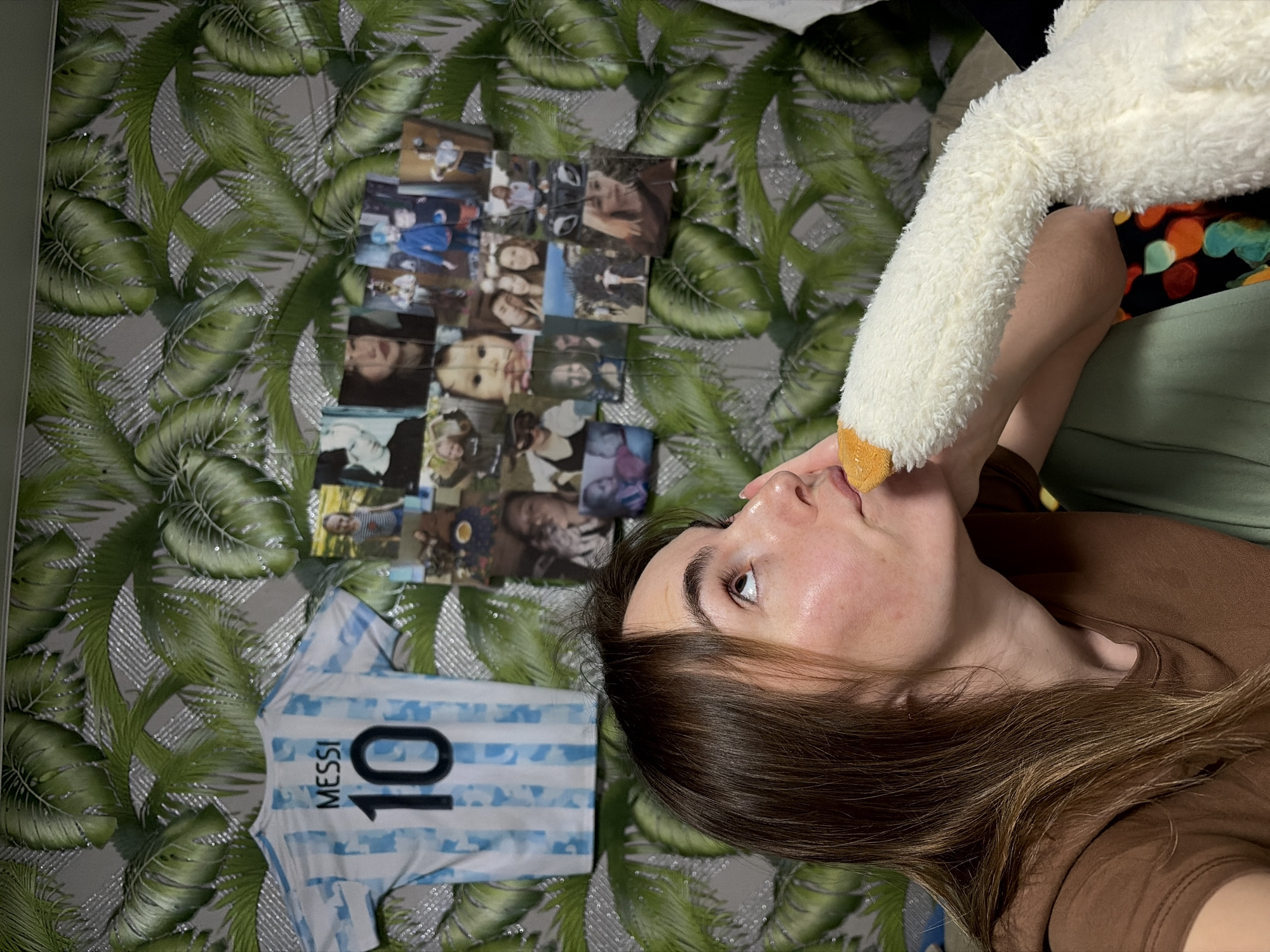 A person with long brown hair, wearing a brown t-shirt and an olive-green shirt, is holding a white stuffed animal goose to their lips. The person is lying on a surface with a green leaf-patterned background. To the left of the person are several printed photographs and a jersey with the number "10" and the name "MESSI" on it. There is no evidence to confirm or deny the location of the image within Nizhnii Vishur, Russia.