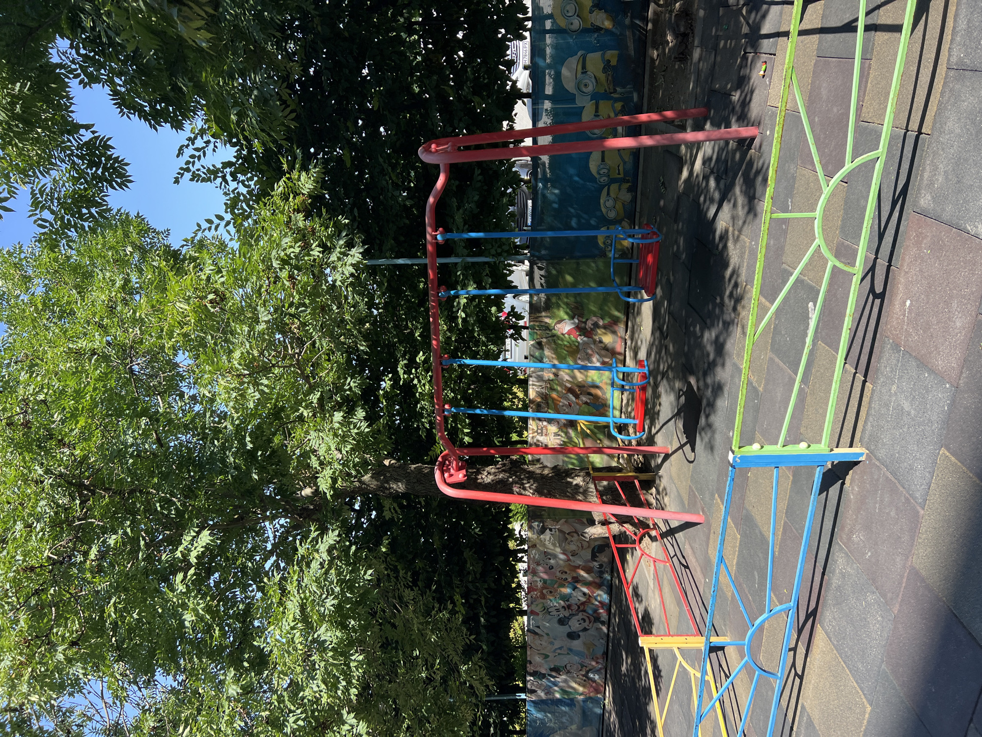 In Fontanka, Ukraine, a children's playground is depicted on a sunny day, with dappled sunlight filtering through the leaves of a large tree in the background. The playground features a red swing set with two blue seats. The ground is covered with a dark-colored tiled surface. A colorful fence with red, yellow, blue, and green sections surrounds the play area. Behind the fence, a mural depicting cartoon characters like Mickey Mouse and the Minions adorns a wall. The environment is outdoors, with lush green foliage and a clear blue sky visible.