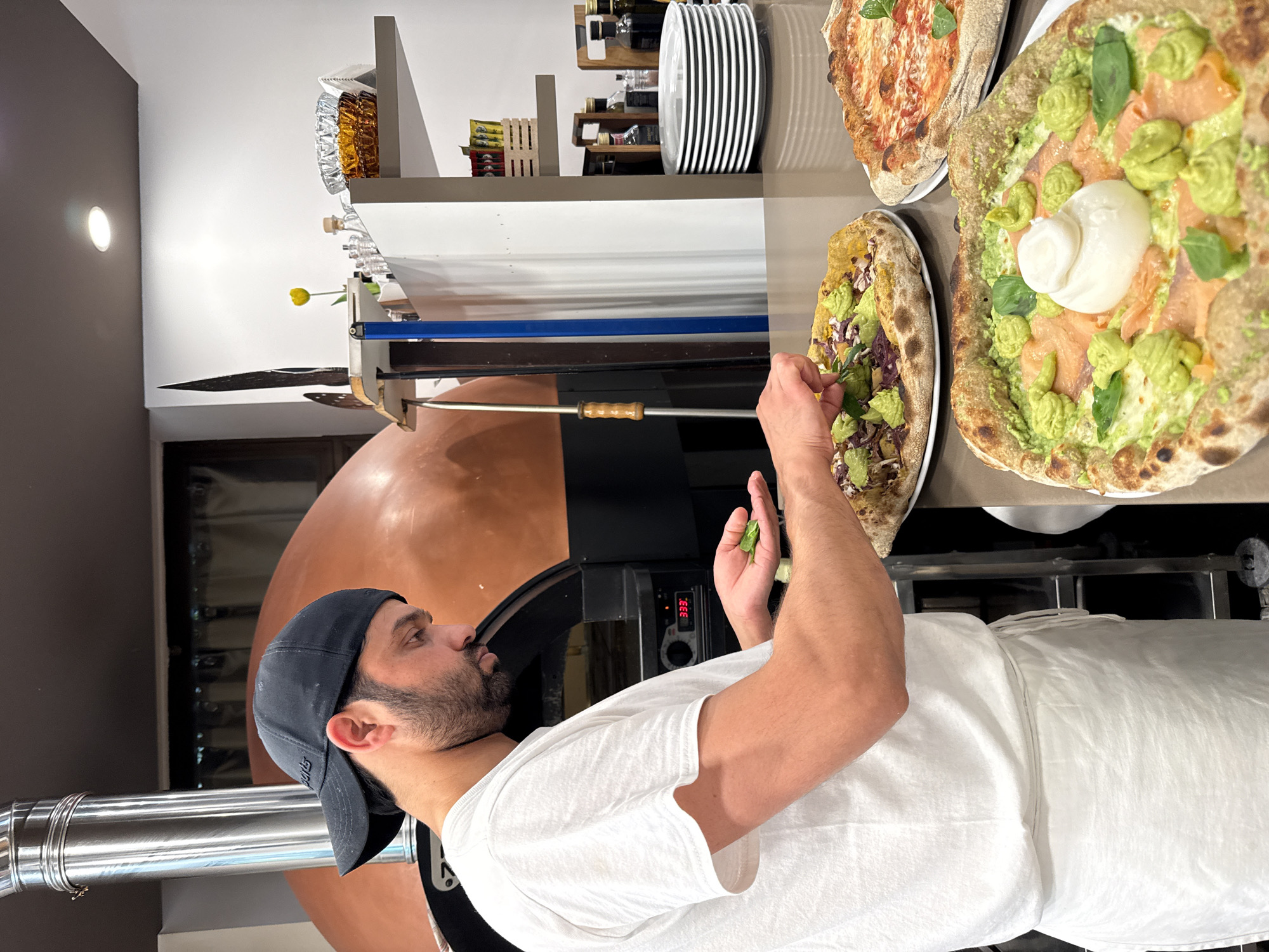 A bearded man wearing a white t-shirt, a black baseball cap, and a white apron is in the process of making a pizza. He stands in front of a large, reddish-brown, dome-shaped pizza oven, which has a stainless steel chimney extending upwards. The man is holding a small green leaf in his left hand and appears to be placing it on a pizza that is already on a metal counter in front of him. This pizza is topped with avocado, red onion, and other ingredients. To the right of this pizza, there are two other pizzas on white plates. One is a margherita pizza with tomato sauce, cheese, and basil. The other pizza, in the foreground, is topped with what appears to be pesto, smoked salmon, fresh basil leaves, and a dollop of burrata cheese. The setting looks like a restaurant or pizzeria kitchen, possibly in Trento, Italy, given the context. Shelves are visible in the background, holding plates, bottles of olive oil, and other kitchen items. A yellow tulip in a small vase is also visible on a shelf, adding a touch of color to the scene. The overall impression is one of fresh food preparation in a professional kitchen.
