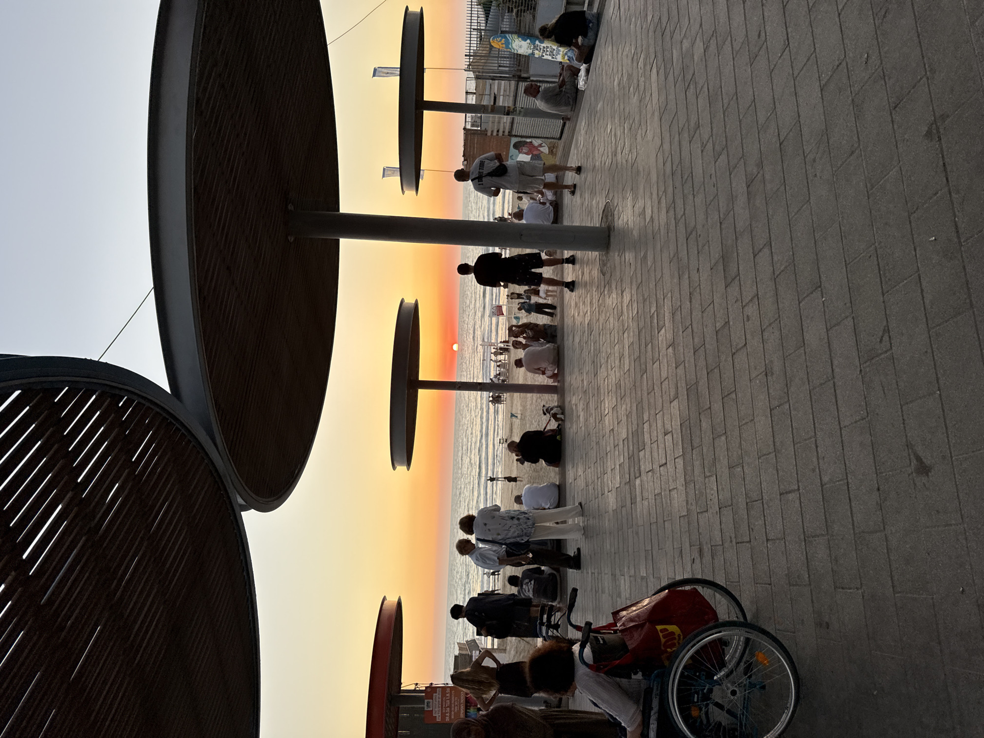 The image captures a sunset scene at the Bat Yam promenade in Israel. The sun is low on the horizon, casting a warm orange glow across the sky and the sea. Several large, circular wooden canopy structures provide shade over a paved walkway.
In the foreground, a woman sits in a wheelchair with a red bag on the back. To her left, another woman in a headscarf stands with her back to the camera. Further back, a group of people are gathered on the paved area, some standing and some sitting.
Beyond the walkway is a sandy beach where more people are dispersed, some near the water's edge. The sea stretches out to the horizon, reflecting the colors of the sunset. In the background, there are some buildings and structures along the coastline. The overall atmosphere is one of relaxation and enjoyment of the coastal evening.