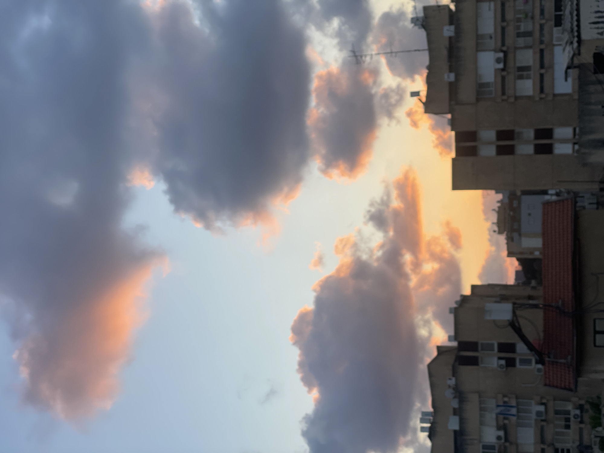 The image depicts a sky with a mix of gray and orange-tinted clouds, suggesting either sunrise or sunset. Tall, multi-story buildings are visible on the right side of the frame, with multiple windows and air conditioning units. The buildings are constructed of beige colored stone and brick with red tiled roofs. The photograph appears to be taken from a slightly lower perspective, looking up at the sky and buildings. This scene is located in the city of Petah Tikva, Israel.