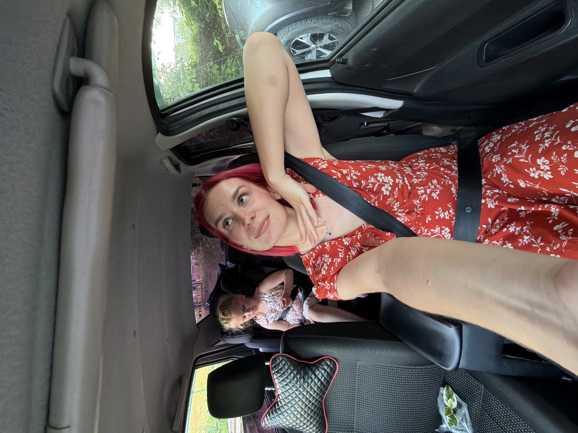 A first-person perspective selfie shows a woman with bright pink hair sitting in the driver's seat of a car. She is wearing a red dress with a white floral pattern and a black seatbelt. Her left arm is resting on the open car door, and her right hand is touching her neck. She is looking up and to her right with a slight smile. In the backseat, a young girl with blonde hair in a pink dress with sequins is seated and appears to be looking down at something in her hands. The interior of the car is visible, including the headrests and seat upholstery. Outside the car window, greenery and another vehicle are visible. The scene appears to be in a residential or suburban area. The location is identified as Chaikovsky, Russia.