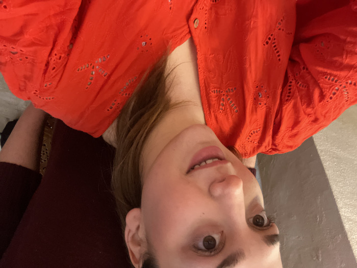 The scene is an indoor location in Ancenis, France. The primary subject is a young female, positioned in the foreground, facing the viewer with an upward and slightly rightward gaze and a subtle smile. She possesses brown eyes and hair, and is attired in a bright red, long-sleeved top featuring eyelet embroidery and a button-front V-neck. A thin gold-tone necklace is present on her neck. Partially visible behind and to the right of the primary subject is another individual, wearing a dark red garment. The background consists of a textured, light-colored wall on the left, with minimal detail visible elsewhere.