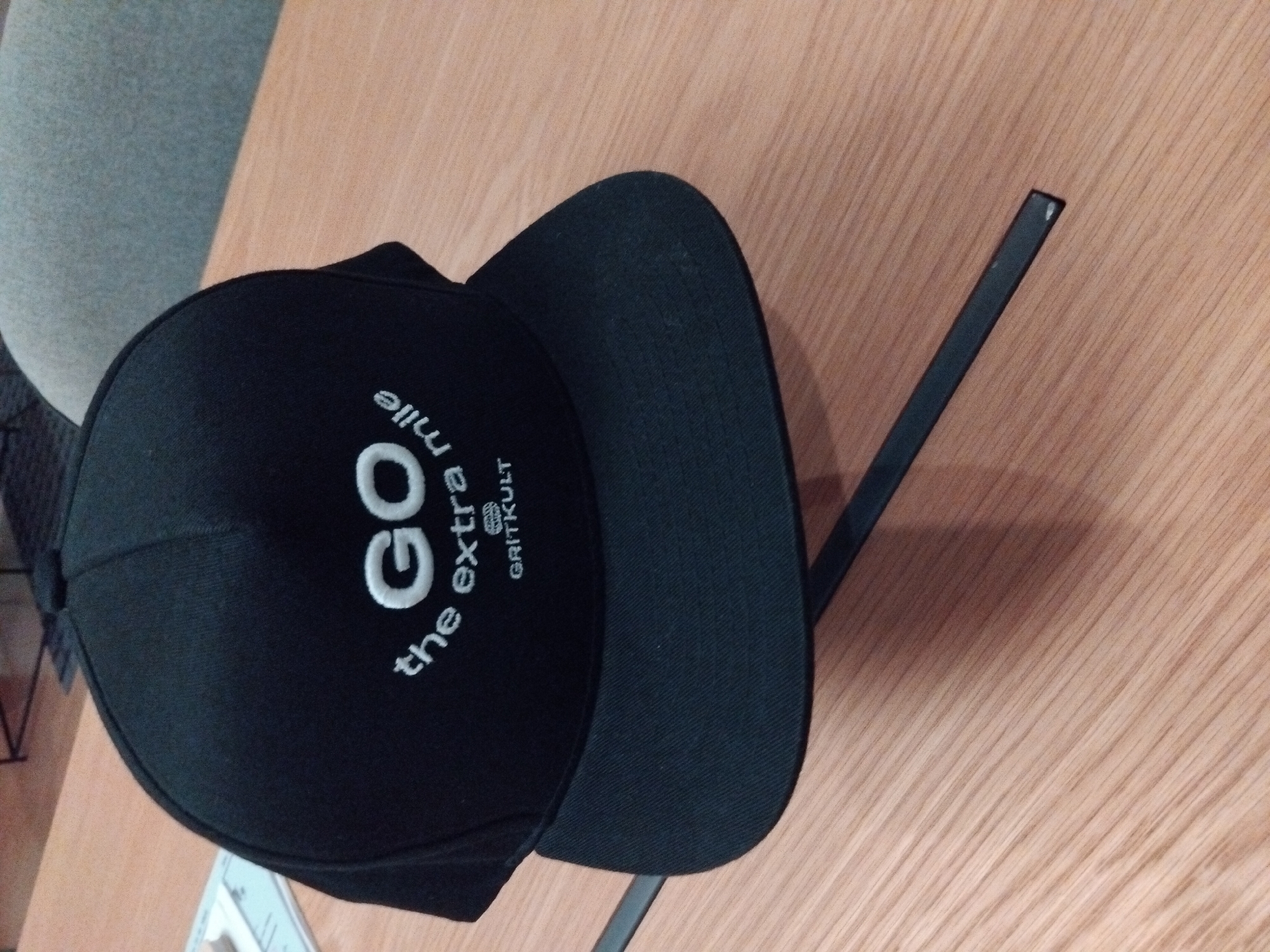 A black snapback cap with white embroidery rests on a wooden table. The embroidery on the cap reads "GO the extra mile" in a stylized font, with the word "GO" being larger. Below this, a smaller embroidery shows the word "GRITKULT" with a circular logo containing a wolf's head. The cap is positioned at an angle, with its brim facing downwards. The table appears to be made of light-colored wood with visible grain. In the background, a portion of a grey chair and a dark metal stand are visible. The scene is indoors. There are no people present. The lighting suggests an indoor environment with ambient light.