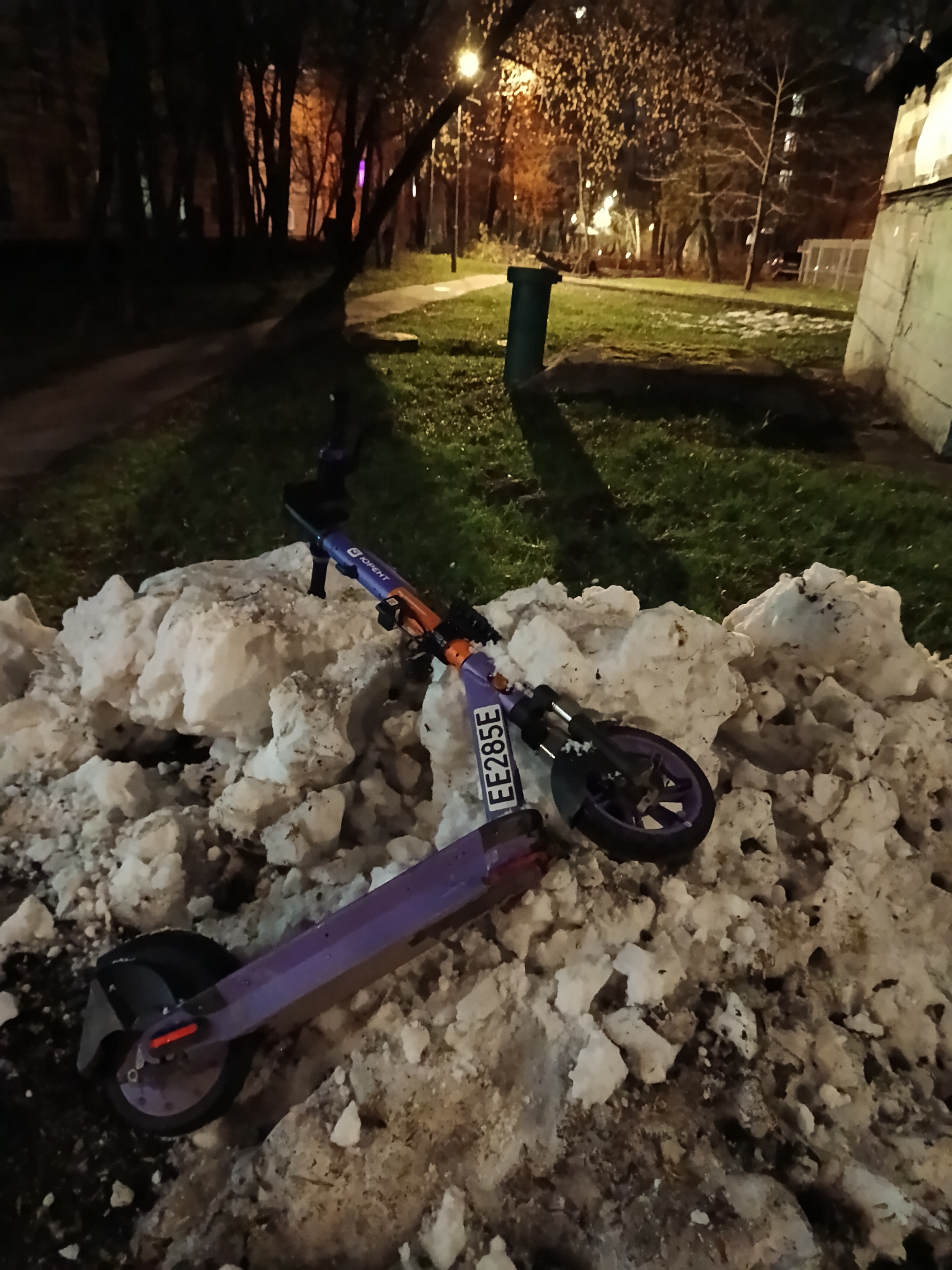 A nighttime photograph captures two abandoned electric scooters resting on a large, dirty pile of melting snow within an urban green space. The scene is illuminated by artificial lights, highlighting patches of green grass and bare trees against the dark sky.
In the foreground, two purple and orange electric scooters are partially buried in the snow. One scooter is more clearly visible, lying on its side with its front wheel and handlebars pointing towards the background. Its stem features the Cyrillic text "ЮРЕНТ" (YURENT), indicative of a scooter rental service. A white plate with the alphanumeric identifier "EE285E" is affixed near its front wheel. The second scooter's rear wheel and part of its platform are visible closer to the bottom left of the frame, also embedded in the snow. The snow pile is uneven, showing signs of dirt and thaw.
Behind the snow, a section of green grass is visible, contrasting with the white and brown snow. A dark green, cylindrical trash can stands on this grassy area towards the right. A paved pathway extends from the left side of the frame, curving into the distance.
The background is dominated by a cluster of bare trees, their dark branches silhouetted against the ambient glow from distant buildings and a prominent streetlamp on the far right. The bright light from the streetlamp casts long shadows and illuminates parts of the park, suggesting a late evening or night setting during a cold season. The visible text "ЮРЕНТ" strongly indicates a Russian location, aligning with the provided context of Moscow, Russia.