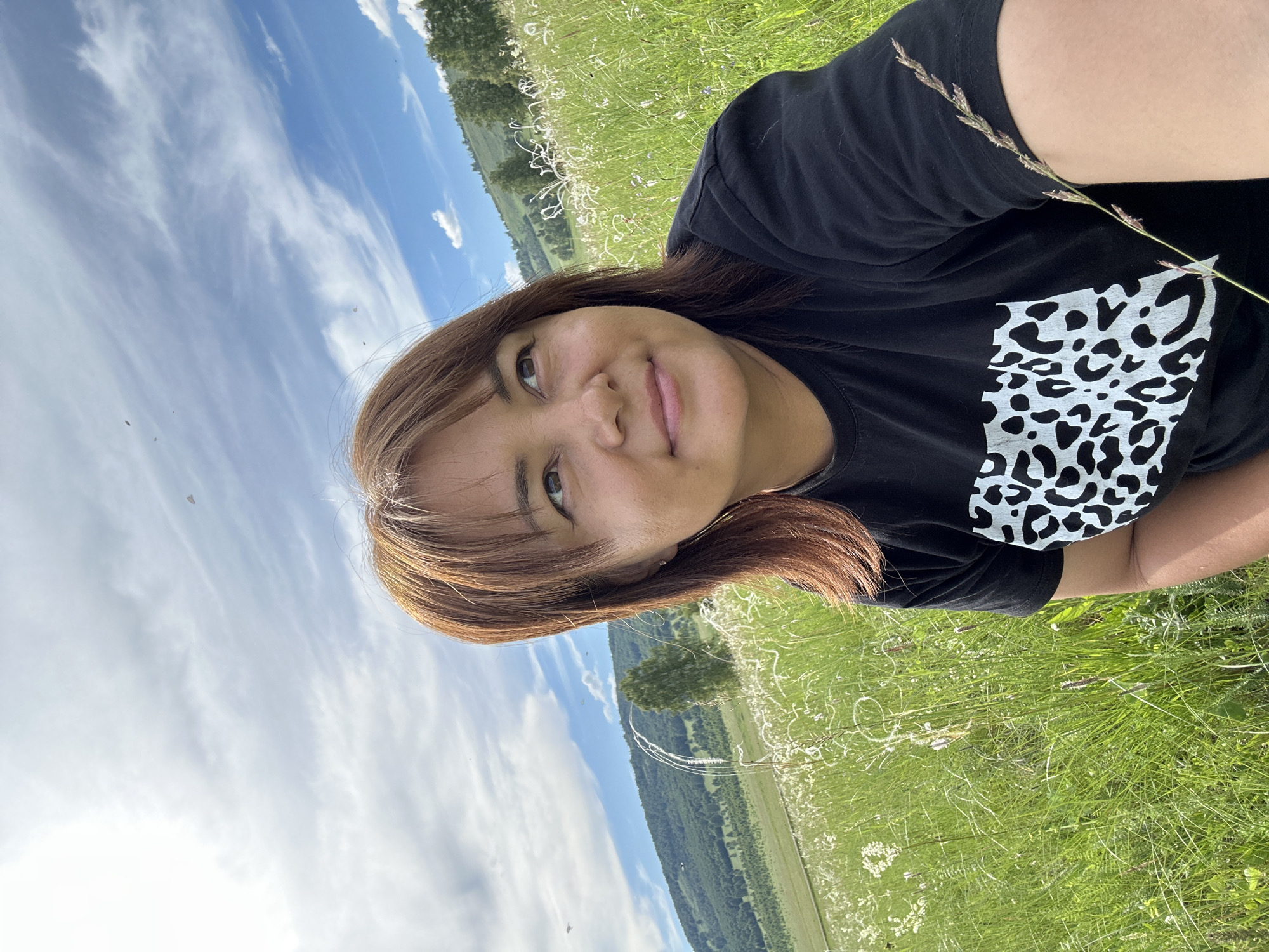 A woman with medium-length brown hair is taking a selfie in a grassy field under a partly cloudy sky. She is wearing a black t-shirt with a white leopard print design on the chest. The background features rolling green hills covered in trees and scattered white wildflowers. A single tall, thin tree stands prominently in the middle ground. The overall scene is natural and serene.
